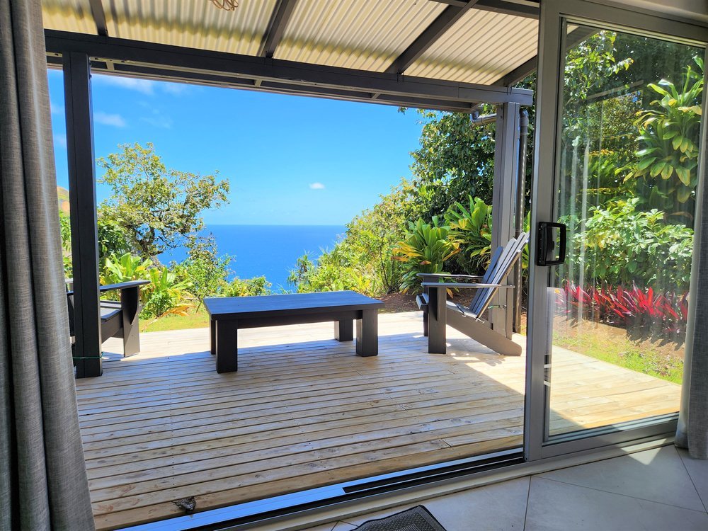 A wooden deck with chairs and a table overlooking a lush garden and ocean view through open sliding glass doors.