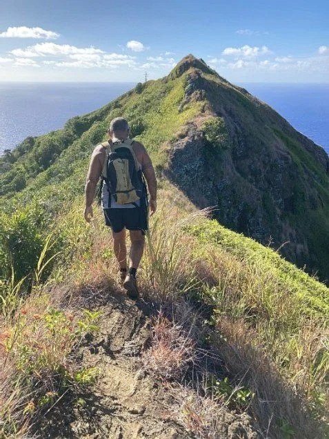 Hiker walking on a narrow trail along a mountain ridge with a steep drop-off on one side, overlooking a large mountain peak and the ocean in the background under a partly cloudy sky.