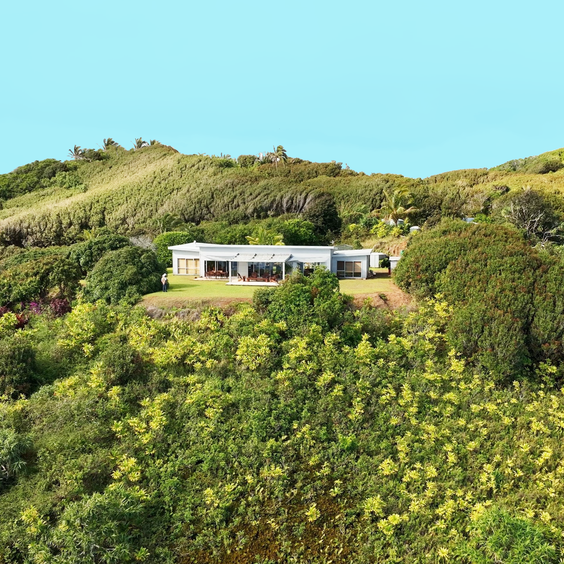 A modern white house surrounded by lush green vegetation and rolling hills under a clear blue sky.