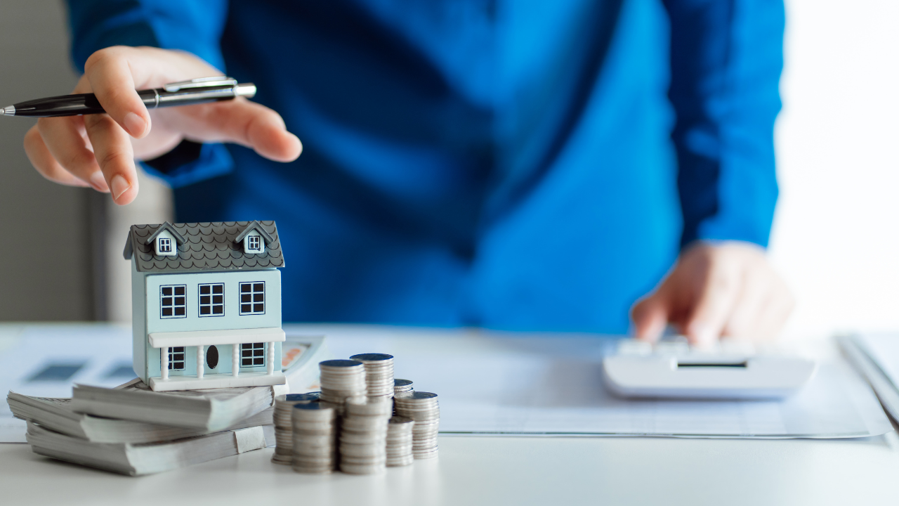 Un uomo in giacca blu che fa una simulazione di compravendita immobiliare con un modellino di casa, monete e documenti su una scrivania