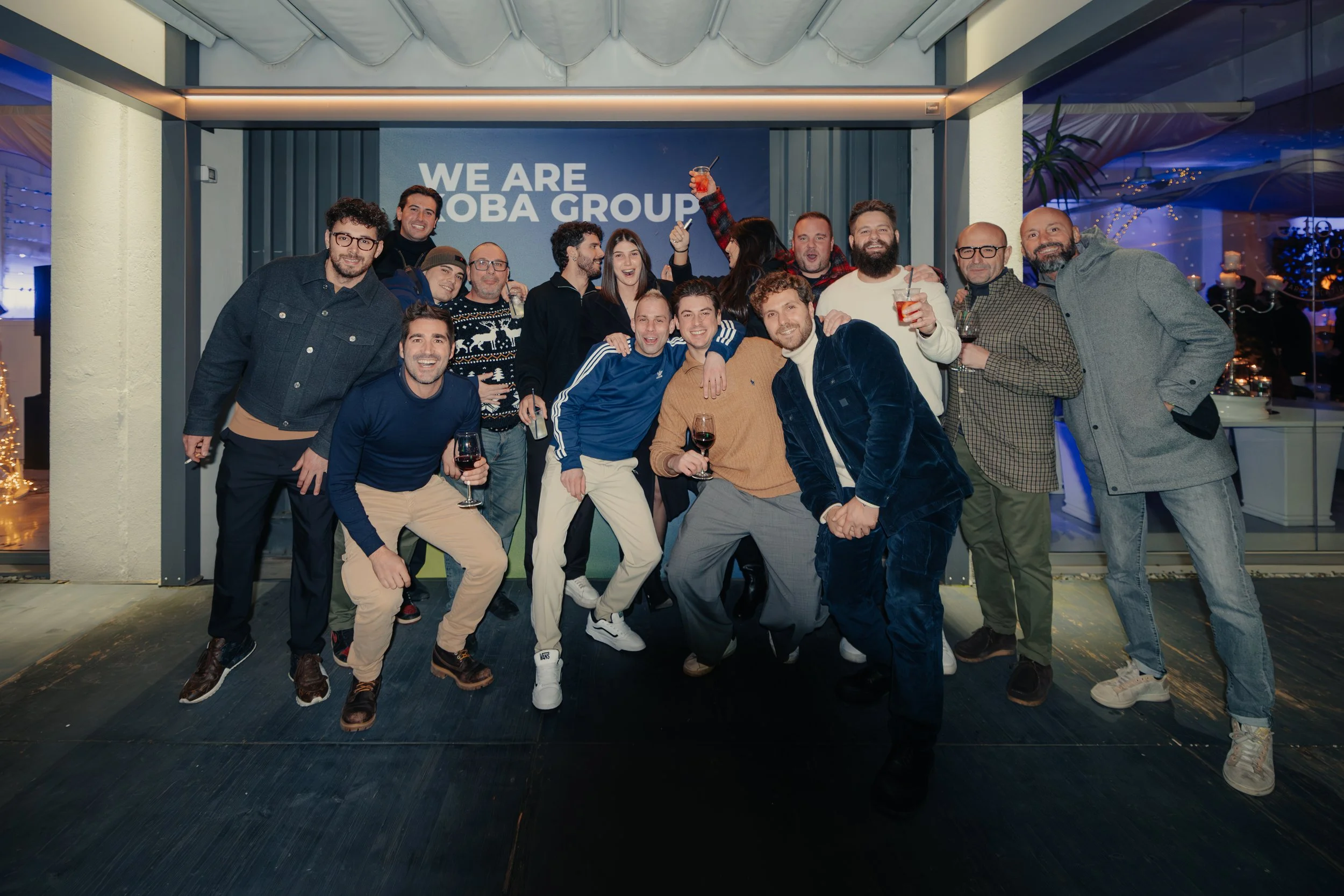 Un gruppo di persone sorridenti e felici che festeggiano con bevande alcoliche, posizionate davanti a una parete con la scritta 'WE ARE TOBA GROUP', all'interno di un locale ben decorato con luci festive.