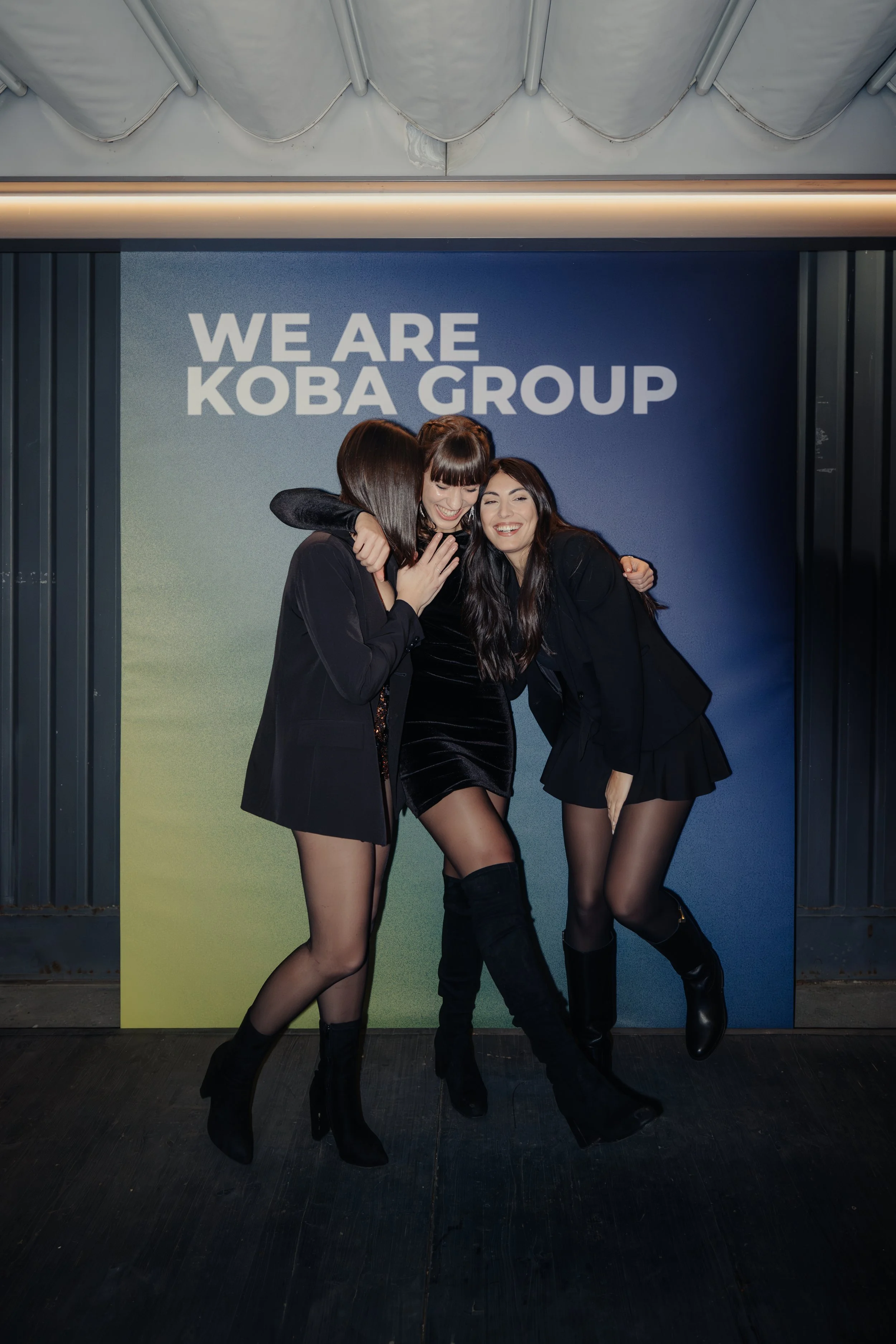 Tre donne in abiti neri che sorridono e si abbracciano davanti a uno sfondo con scritto 'WE ARE KOBA GROUP'.