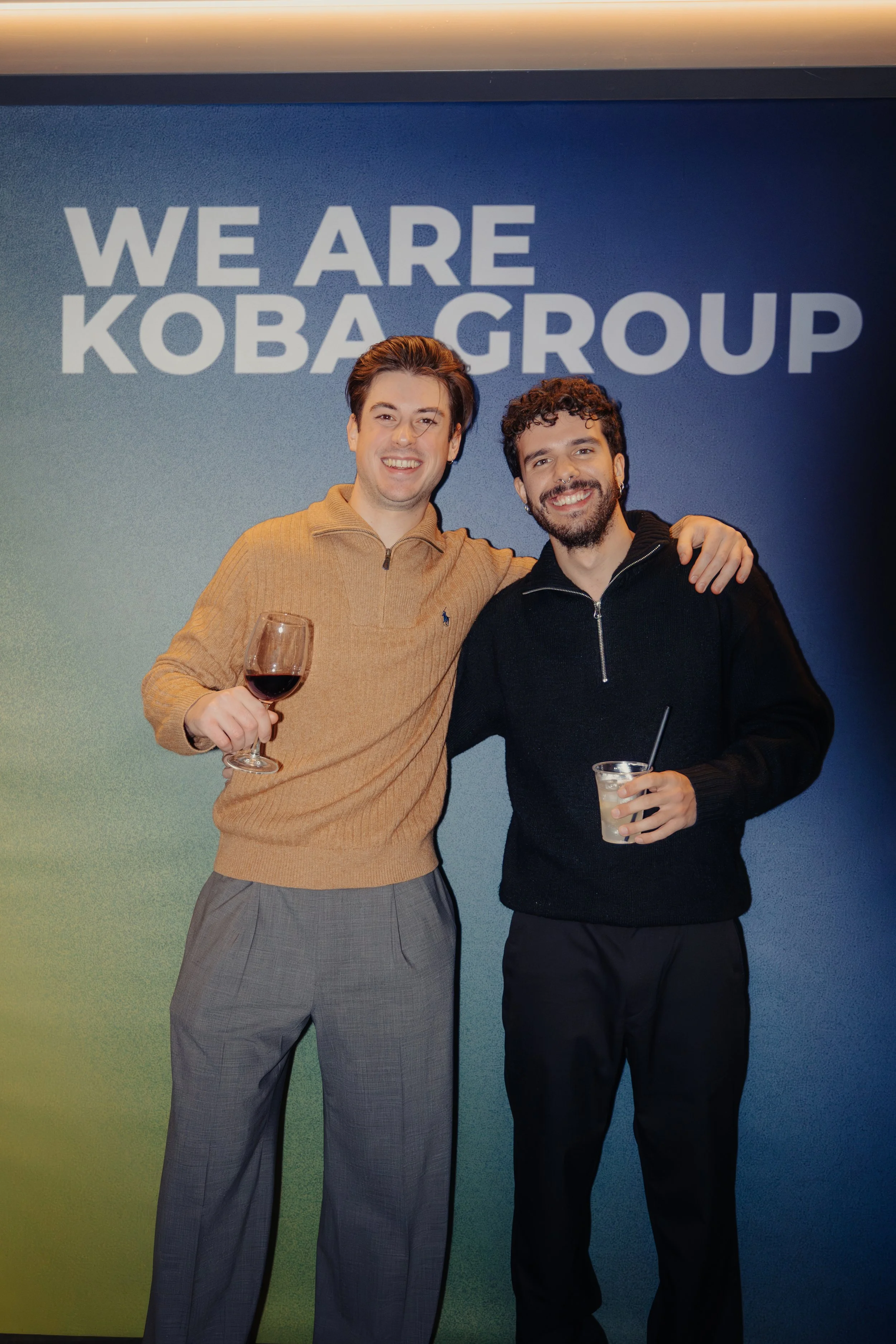 Due uomini sorridenti in piedi davanti a uno sfondo con la scritta 'We Are Koba Group'. Uno tiene un bicchiere di vino rosso e l'altro una bevanda con una cannuccia.