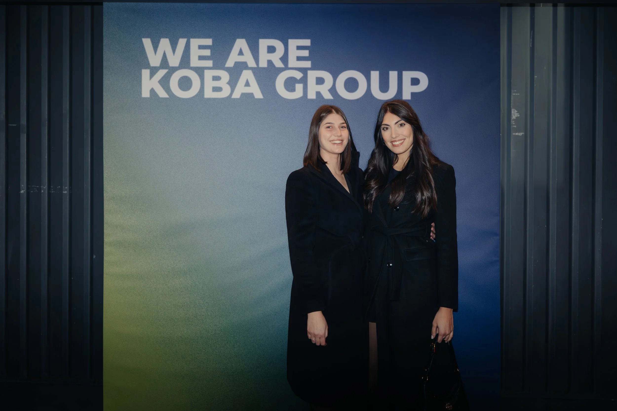 Due donne in abiti neri sorridenti davanti a un muro blu con la scritta 'WE ARE KOBA GROUP'.