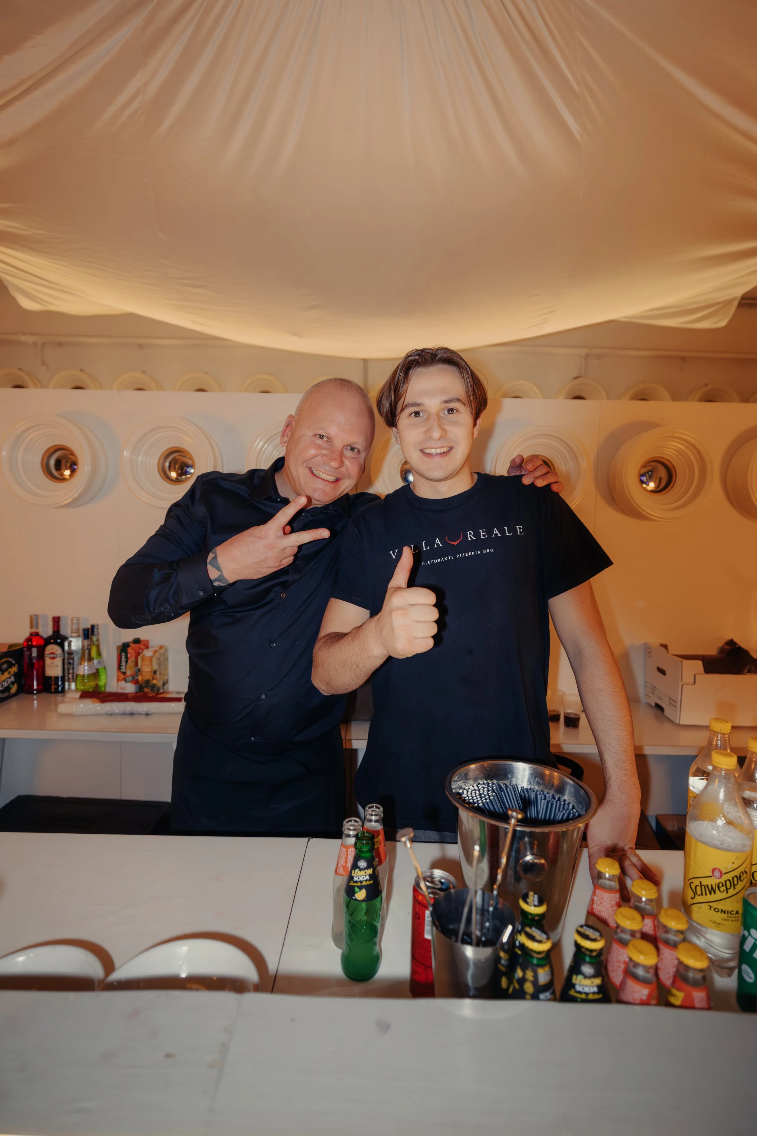 Due uomini sorridenti in un bar con bottiglie e bicchieri alle spalle, uno dei quali indossa una maglietta con scritto Villa Reale, mentre fanno un gesto di approvazione e salute.
