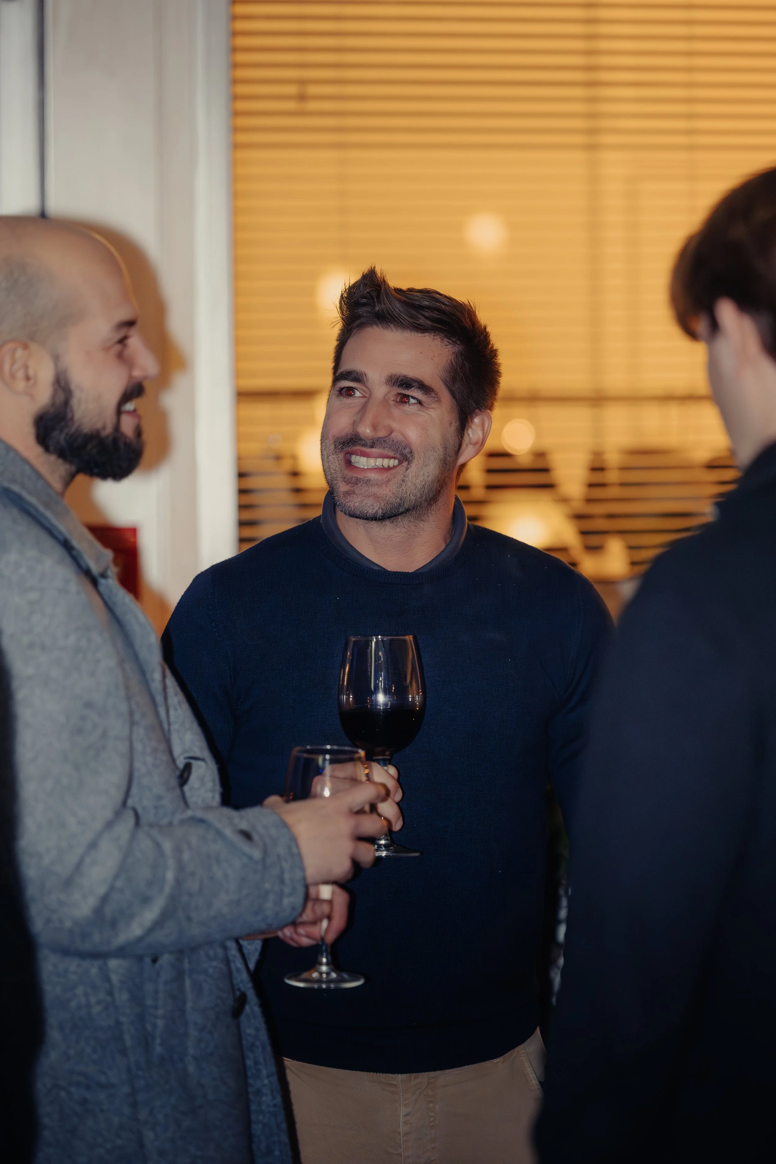 Tre uomini che ridono e parlano durante una conversazione a una festa, uno di loro tiene un bicchiere di vino rosso.