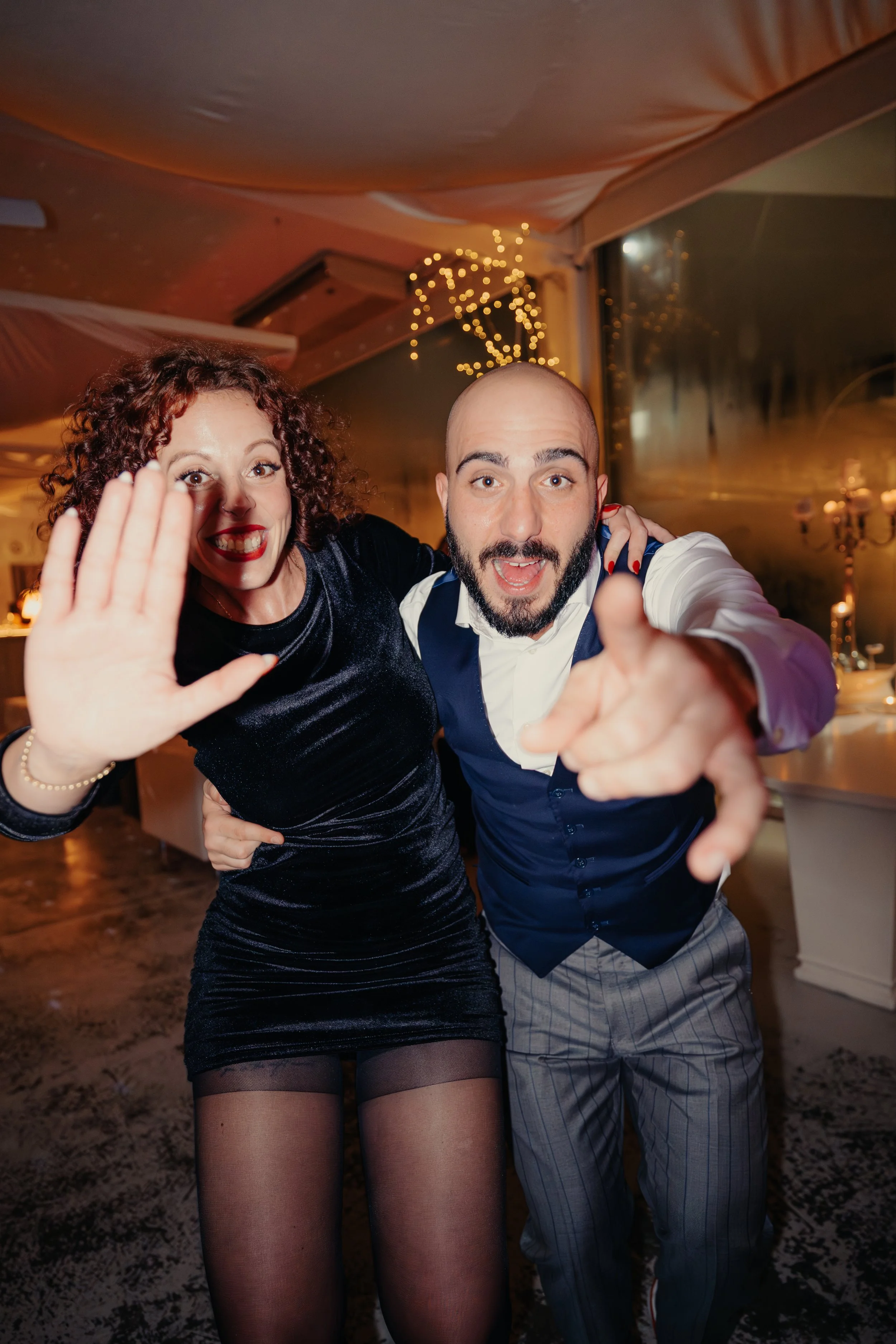 Due persone che sorridono e fanno un gesto di saluto o invito verso la fotocamera, in un ambiente elegante con decorazioni luminose e un'atmosfera festosa.