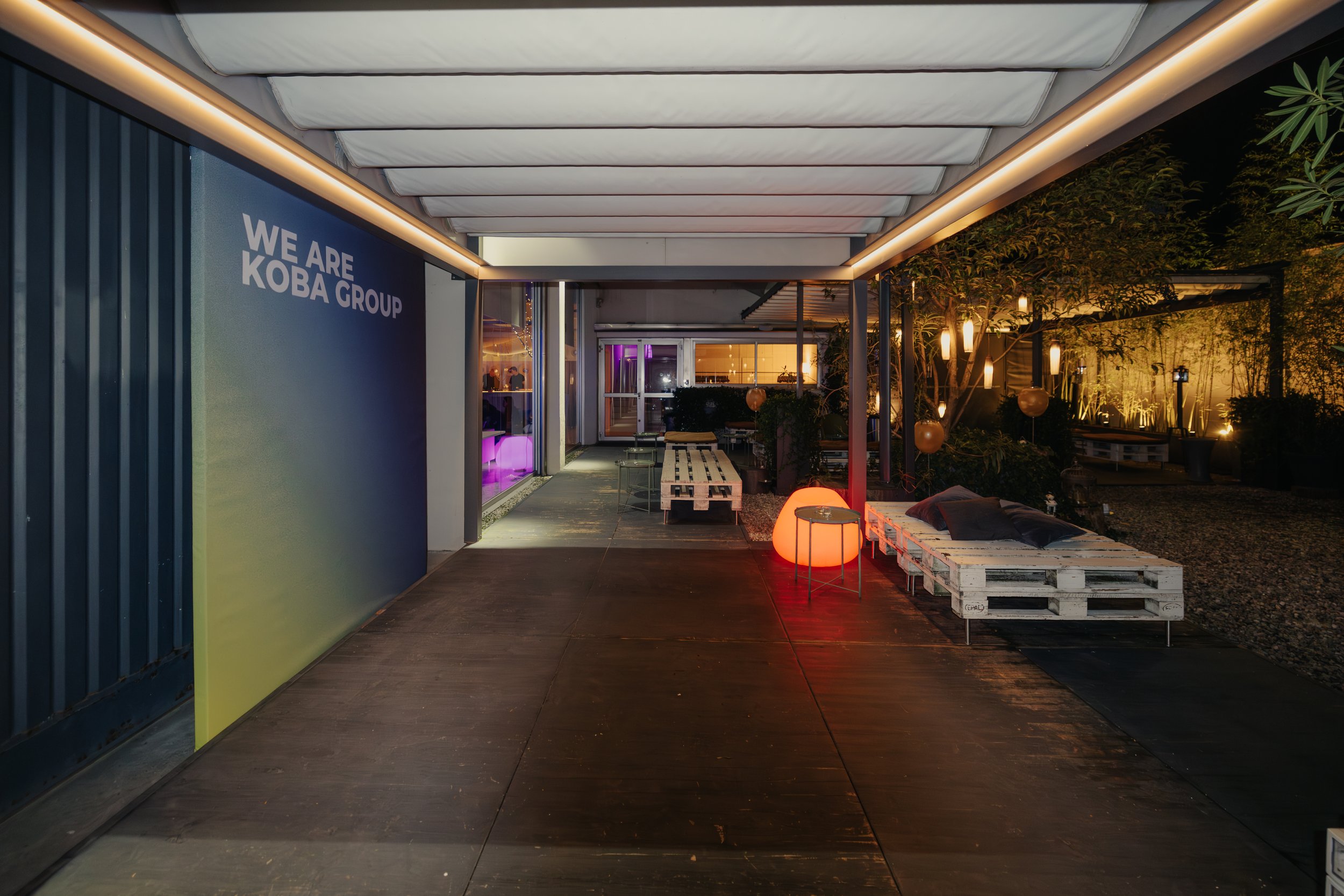 Ingresso di un locale notturno con decorazioni luminose, sedute di pallet e una grande parete con la scritta 'WE ARE KOBA GROUP'.
