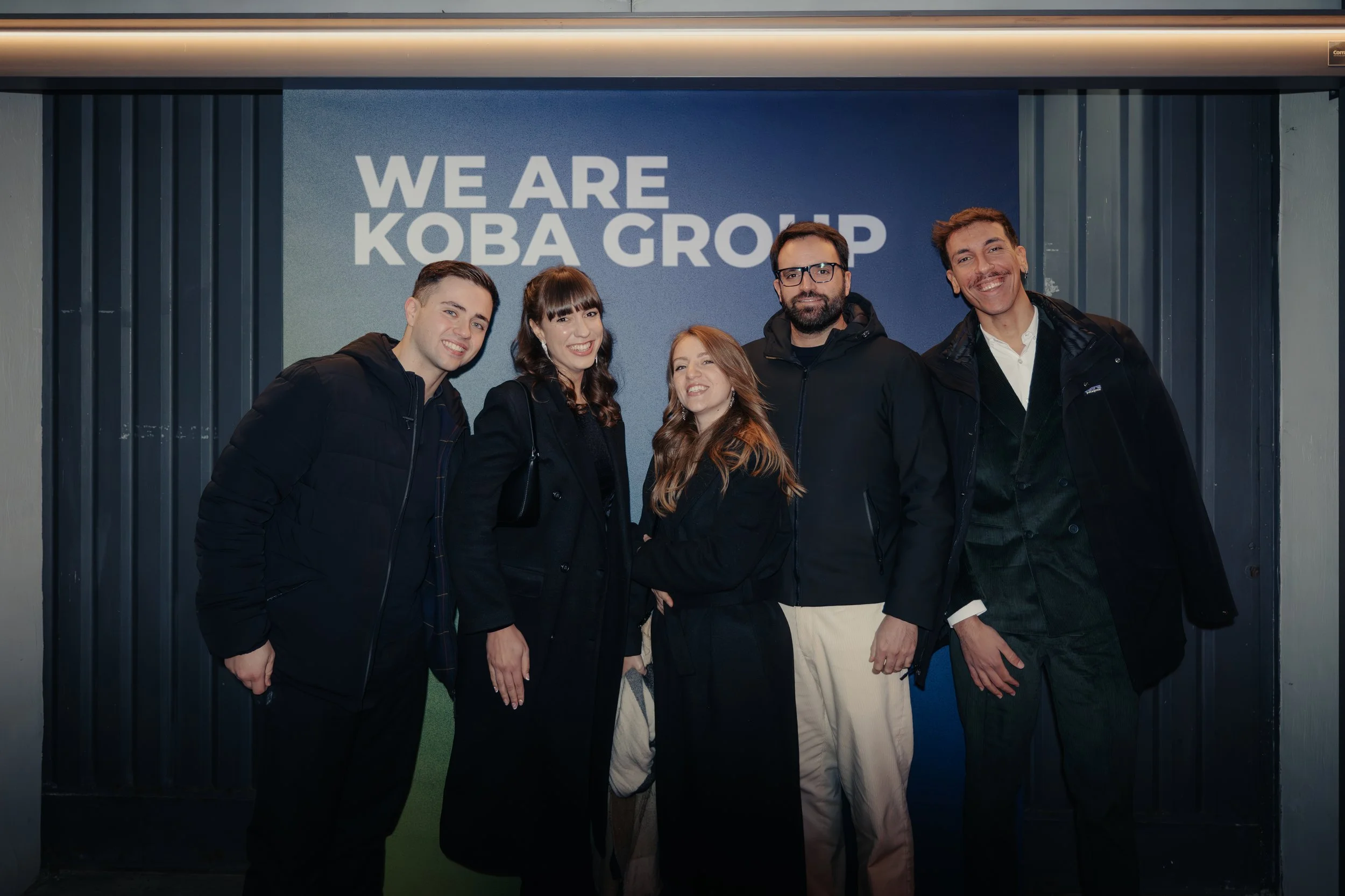 Gruppo di cinque persone sorridenti davanti a un cartellone con scritta 'WE ARE KOBA GROUP'.