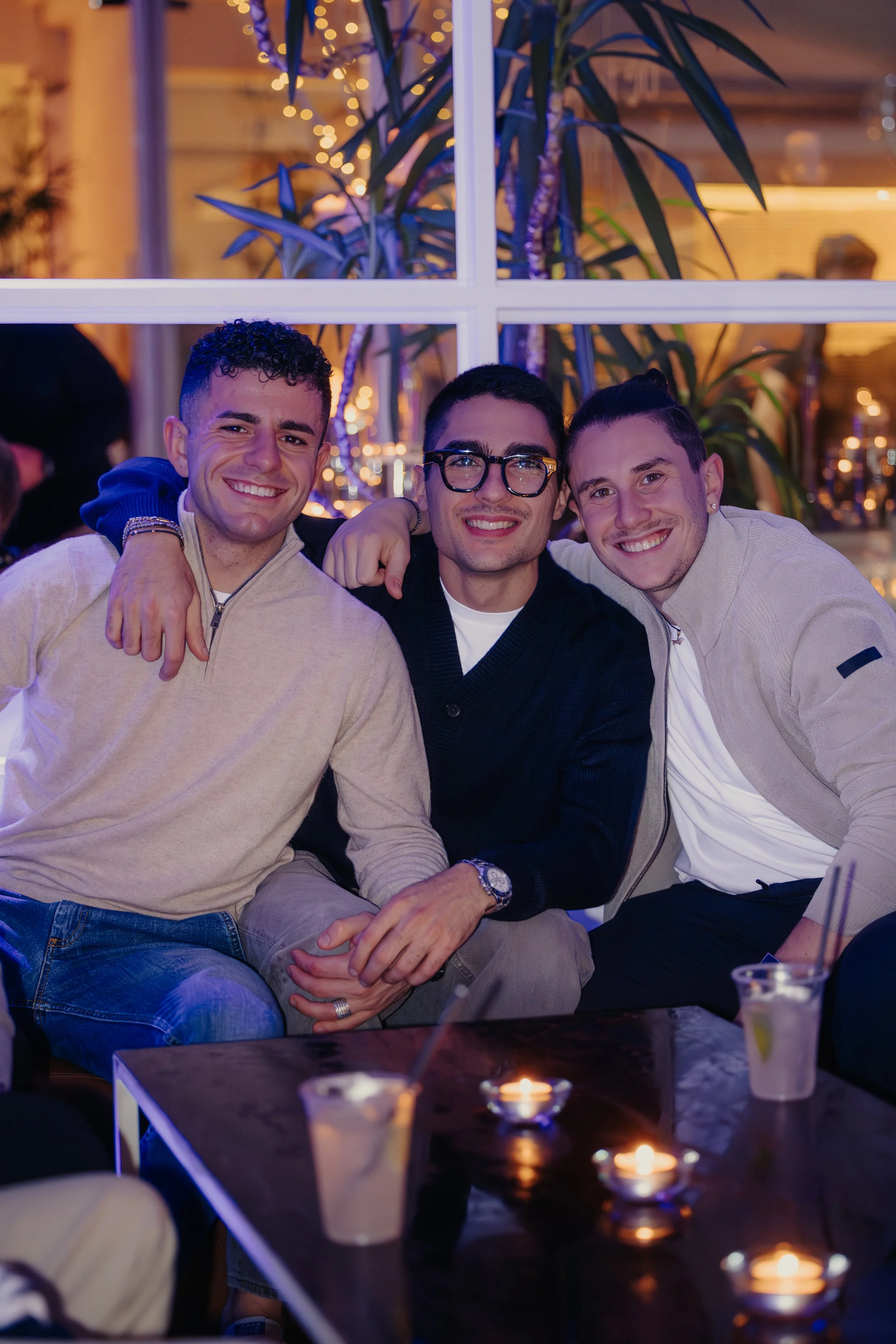 Tre uomini sorridenti seduti insieme in un locale con decorazioni luminose alle spalle.