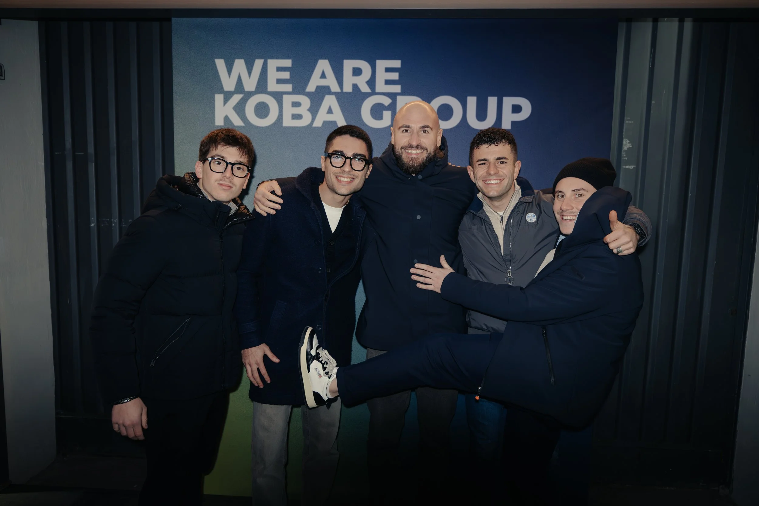 Gruppo di cinque uomini sorridenti e abbracciati davanti a un poster con scritto 'WE ARE KOBA GROUP' in un ambiente al coperto.