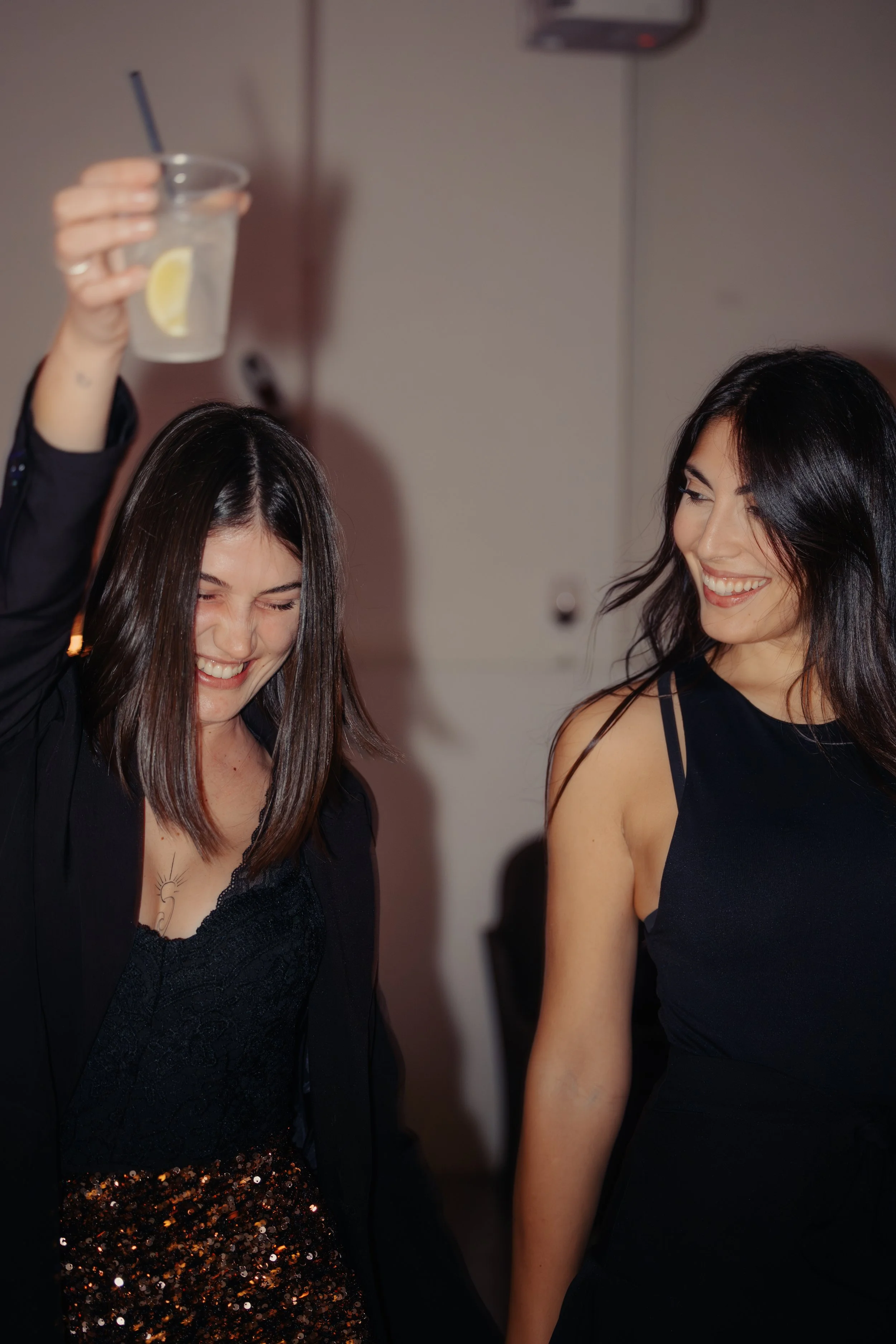 Due donne che ridono e si divertono, una con una bevanda, in un ambiente festoso.