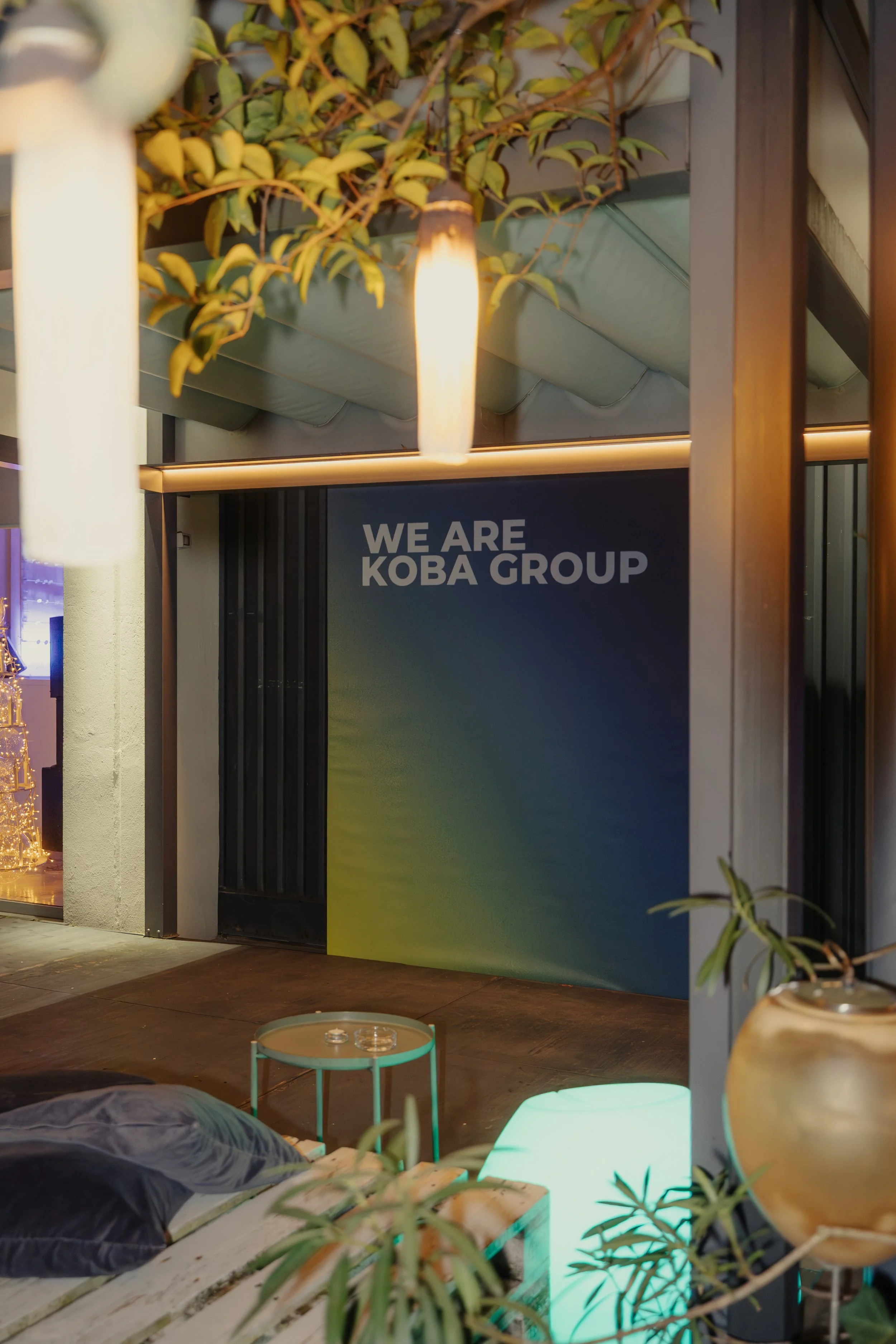 Segnale luminoso con le scritte "WE ARE KOBA GROUP" in un ambiente moderno con piante e luci decorative.