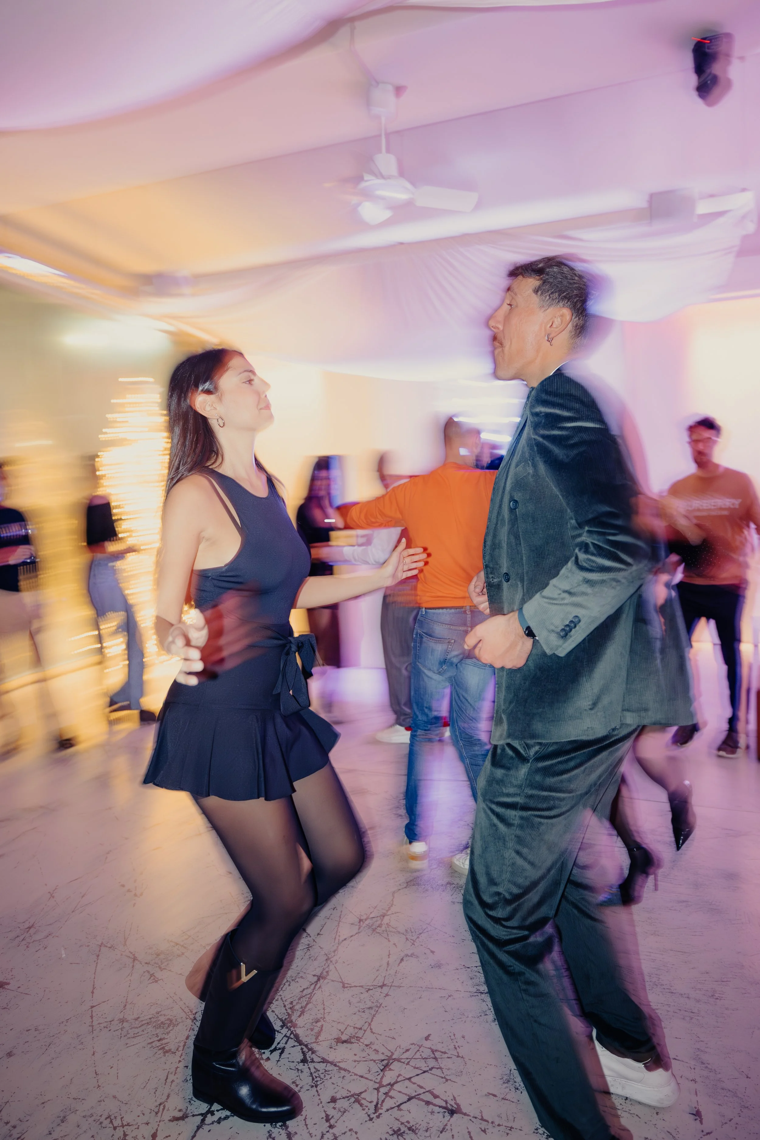Due persone che ballano insieme in una discoteca con luci colorate. La donna indossa un vestito nero e le scarpe nere, l'uomo indossa un abito scuro. Sullo sfondo ci sono altre persone che ballano e decorazioni luminose.