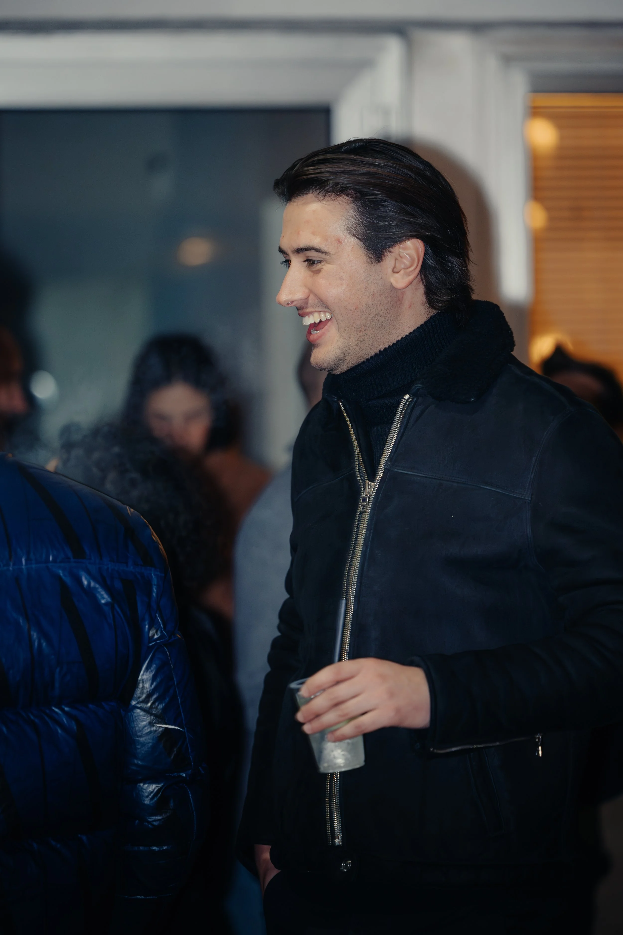 Un uomo sorridente in abbigliamento nero si trova in un ambiente sociale, tenendo un bicchiere in mano, circondato da altre persone.