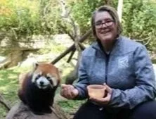 Woman with glasses smiling and sitting outdoors next to a red panda in a wooded area.