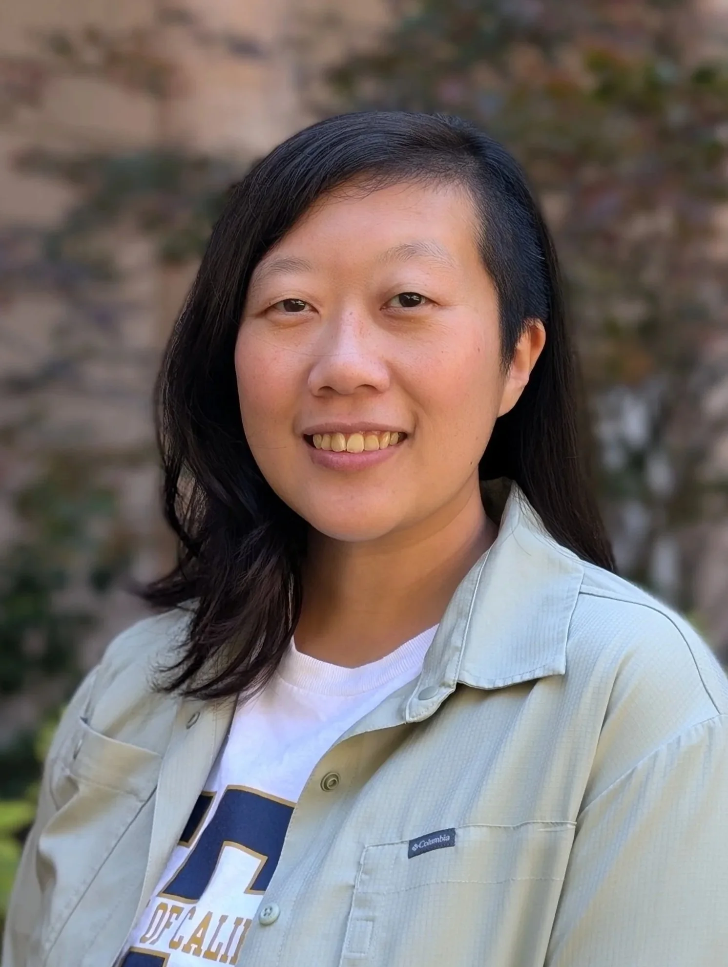 A woman with medium-length black hair, wearing a white T-shirt and a light beige Columbia jacket, smiling outdoors with a blurred natural background.