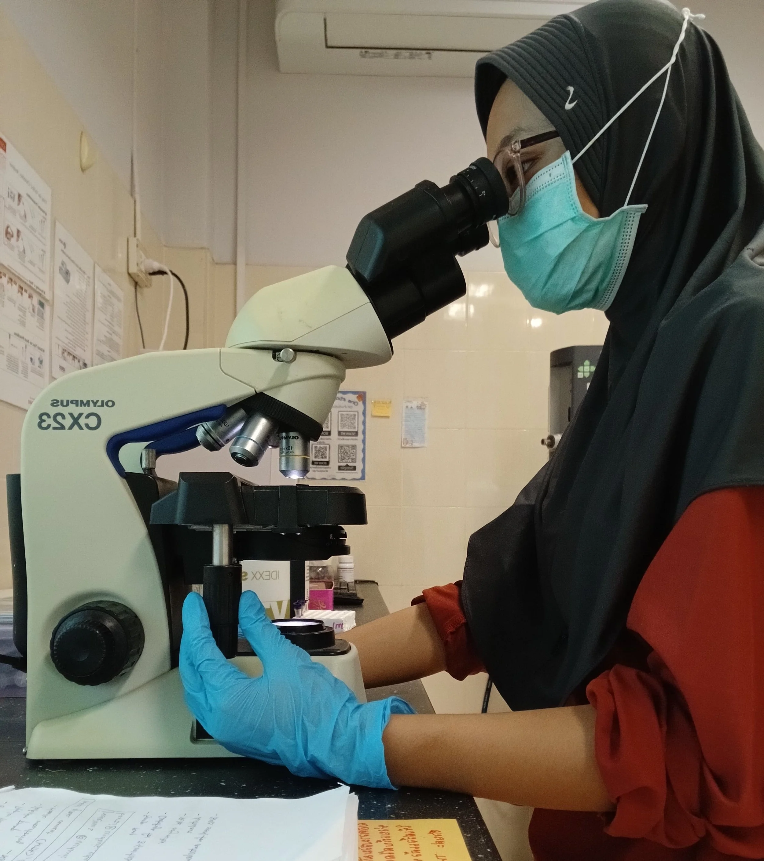 A woman wearing a hijab, mask, and gloves looking through a microscope in a laboratory.