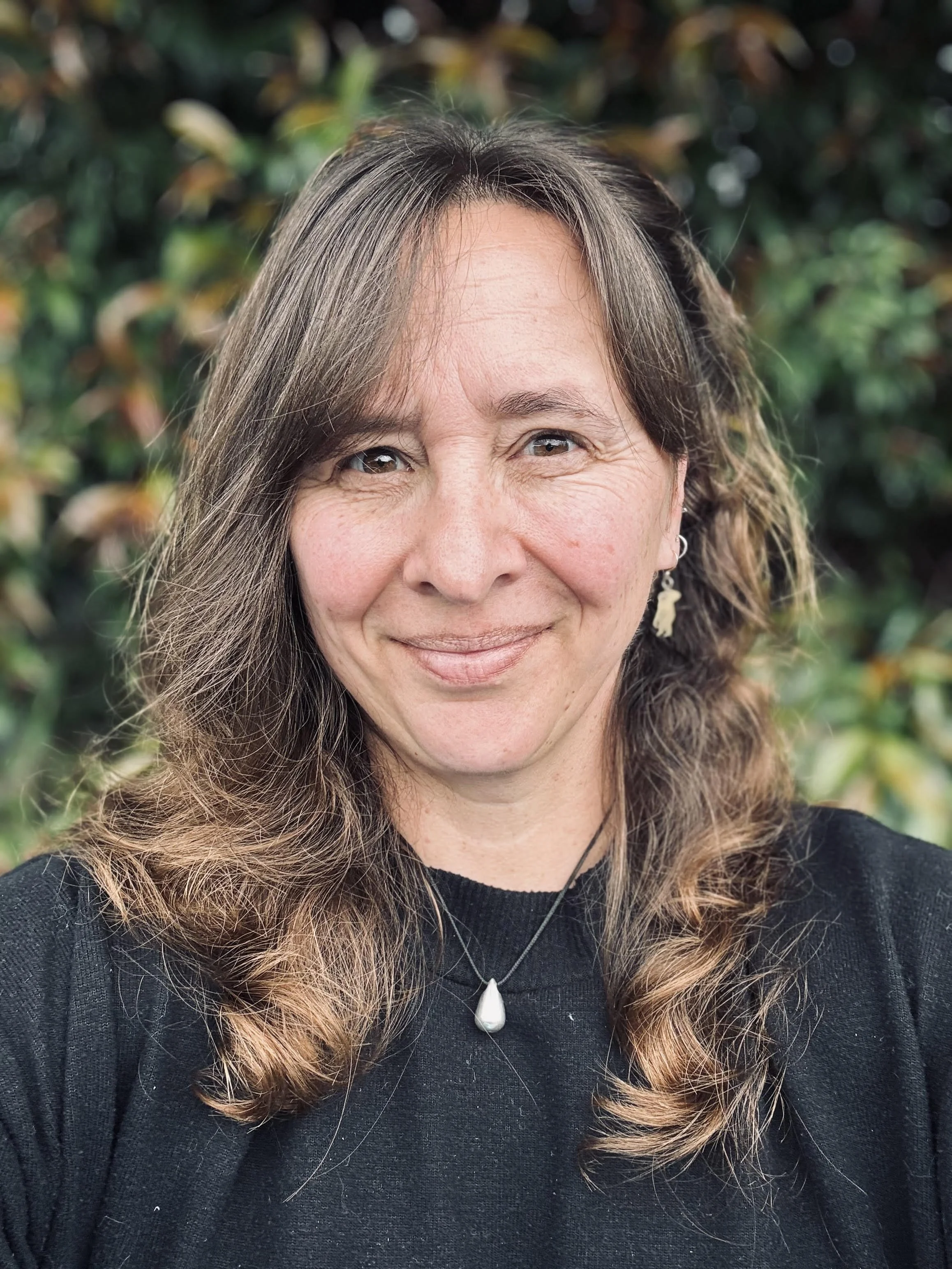 A woman smiling outdoors with a background of green foliage, wearing a black top, a necklace, and earrings.