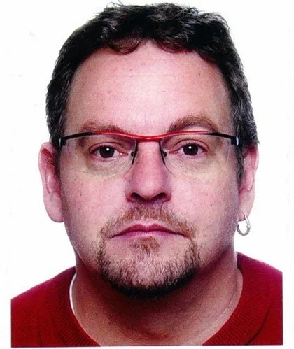 Close-up of a man with glasses and a goatee, wearing a red sweater and a small earring in his left ear.