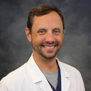 Portrait of a man with brown hair and a beard, wearing a white lab coat, smiling against a dark background.