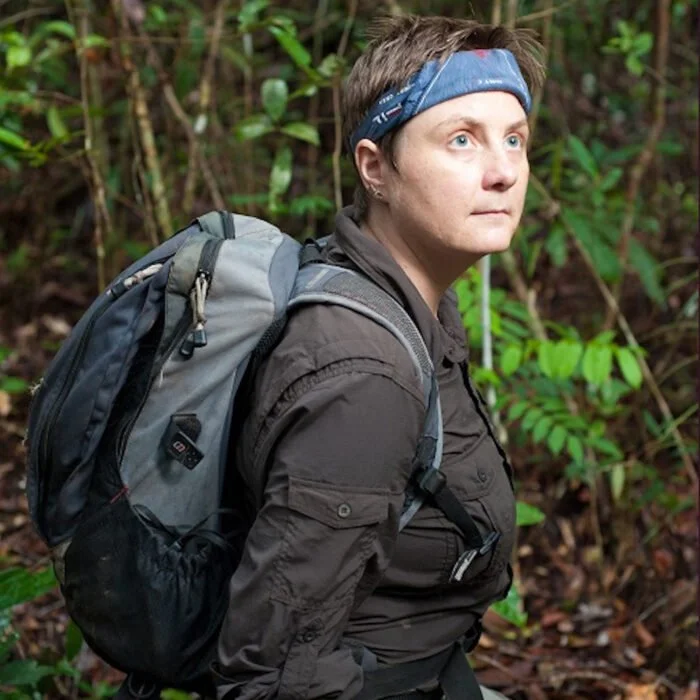 Woman with short hair and a blue headband carrying a gray backpack, standing in a lush green forest.
