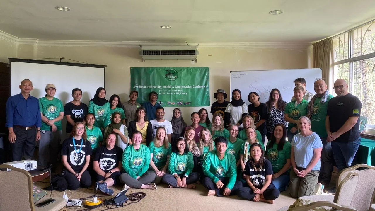 Group photo of people attending the 4th Gibbon Husbandry, Health & Conservation Conference in Palang, with some wearing green conference shirts, standing and sitting in a room with a banner, whiteboard, and window with curtains.