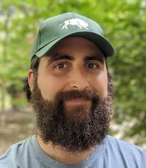 A man with a beard wearing a green cap outdoors, with green foliage in the background.