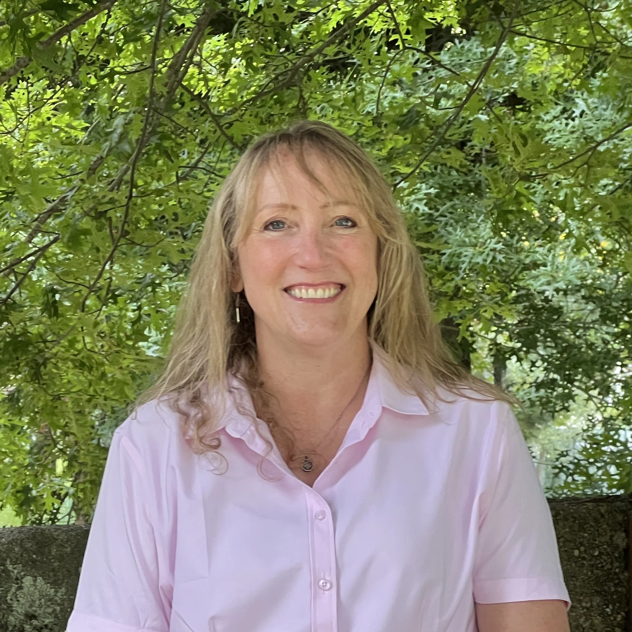 A woman with blonde hair smiling outdoors in front of green leafy trees, wearing a light pink button-up shirt and a necklace.