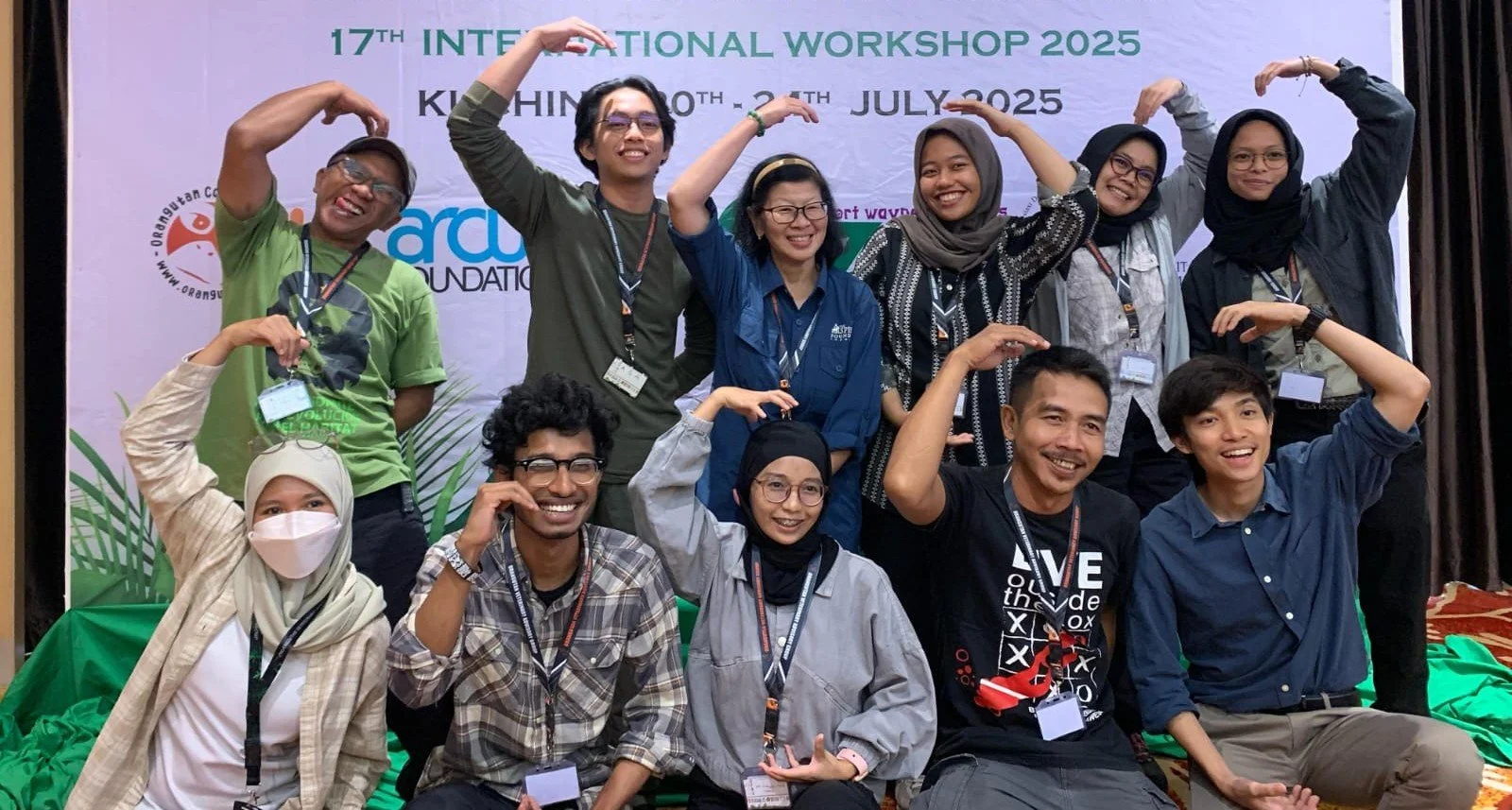 A group of people posing on stage at an international workshop, smiling and making arm gestures. The background features a banner with event details. The group includes diverse men and women, some wearing glasses and masks.