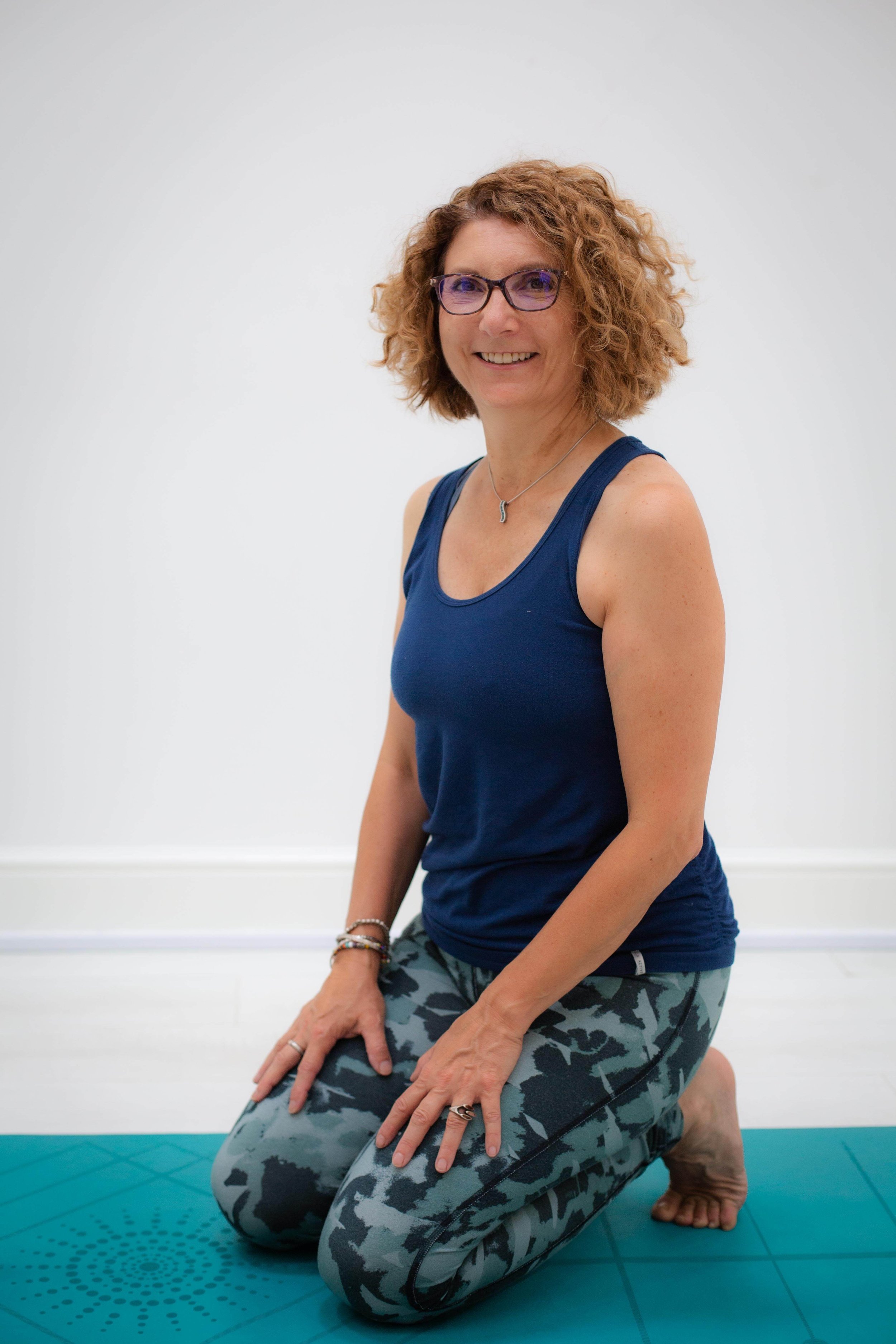 Woman with curly hair wearing glasses kneeling on a yoga mat indoors.