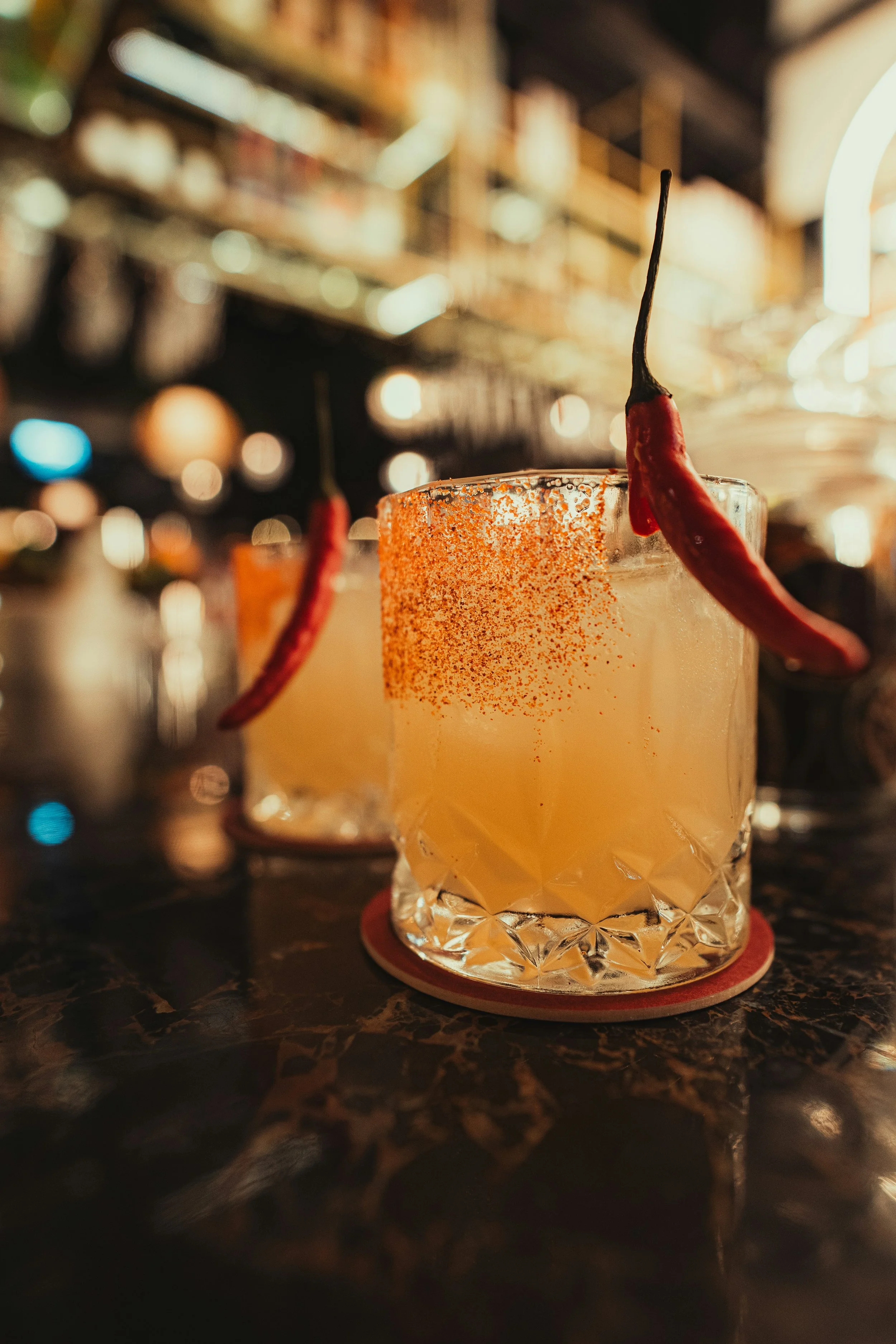 Close-up of a cocktail garnished with a red chili pepper, served in a glass with a salted rim, on a dark marble bar counter in a dimly lit bar or restaurant.