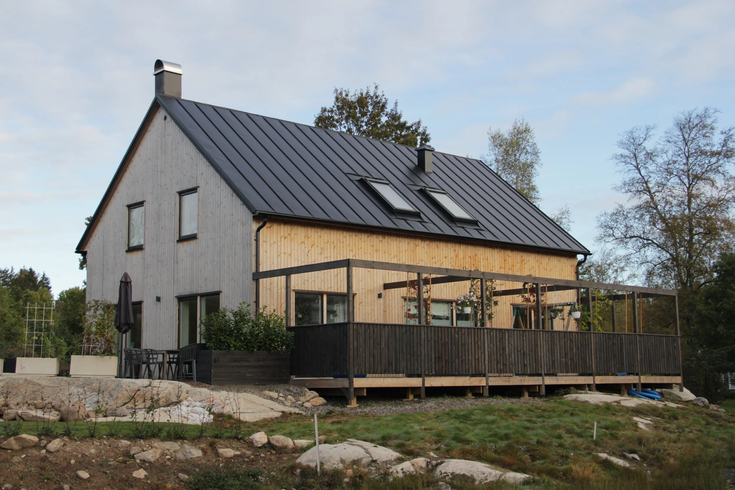 Modernt trähus med svart tak och altan, omgiven av träd.