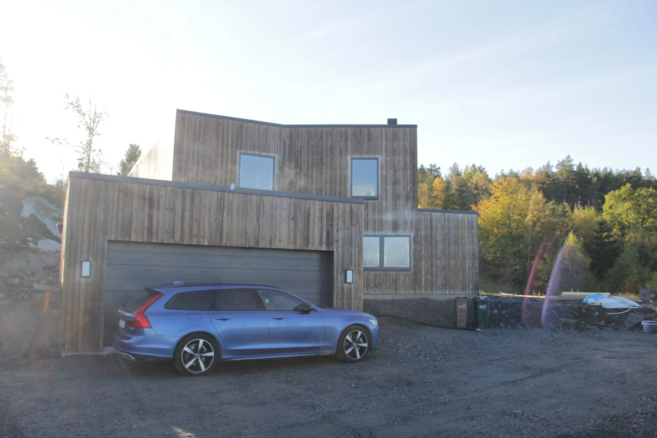 En modern träklädd byggnad med en blå bil parkerad framför. I bakgrunden syns skog.