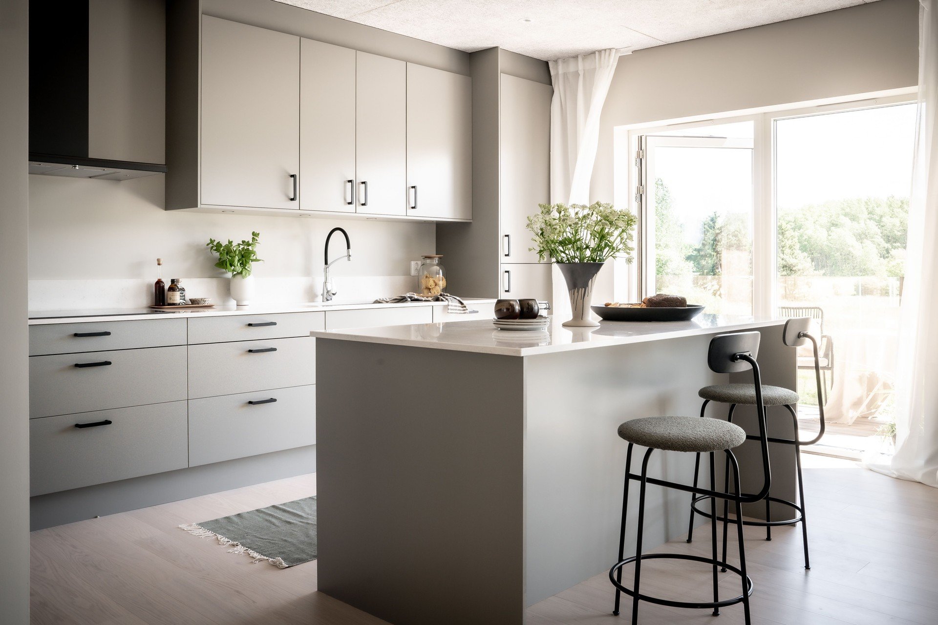 Modernt kök med ljusa skåp, köksö, barstolar, blommor på bordet, stora fönster med utsikt över grönska. O Lind AB.