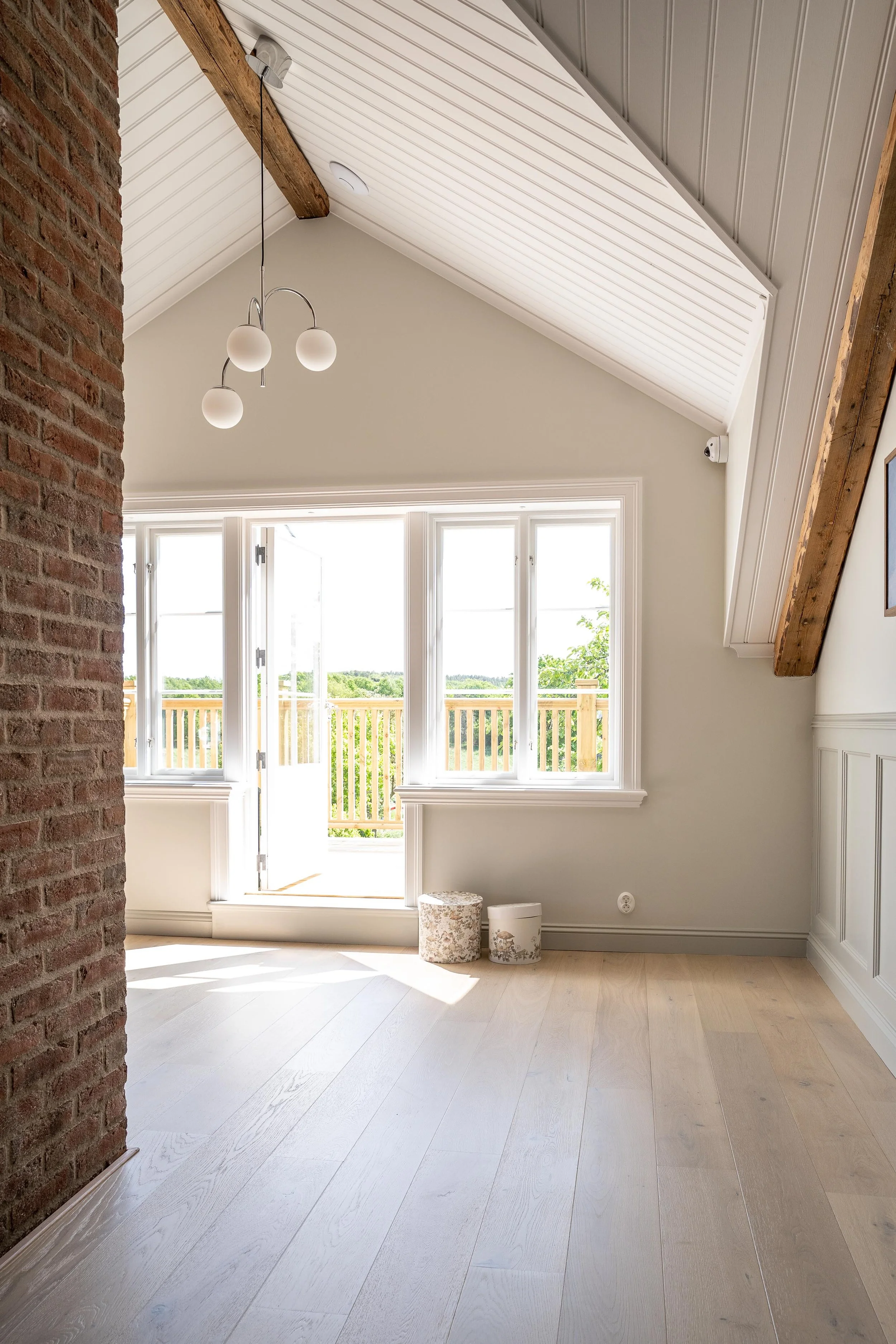Ljust rum med högt i tak, synliga träbalkar och tegelvägg. Stort fönster med utsikt mot veranda och grönska. Ljus trägolv och modern hängande lampa. Mjuka dekorativa föremål på golvet. O Lind AB.