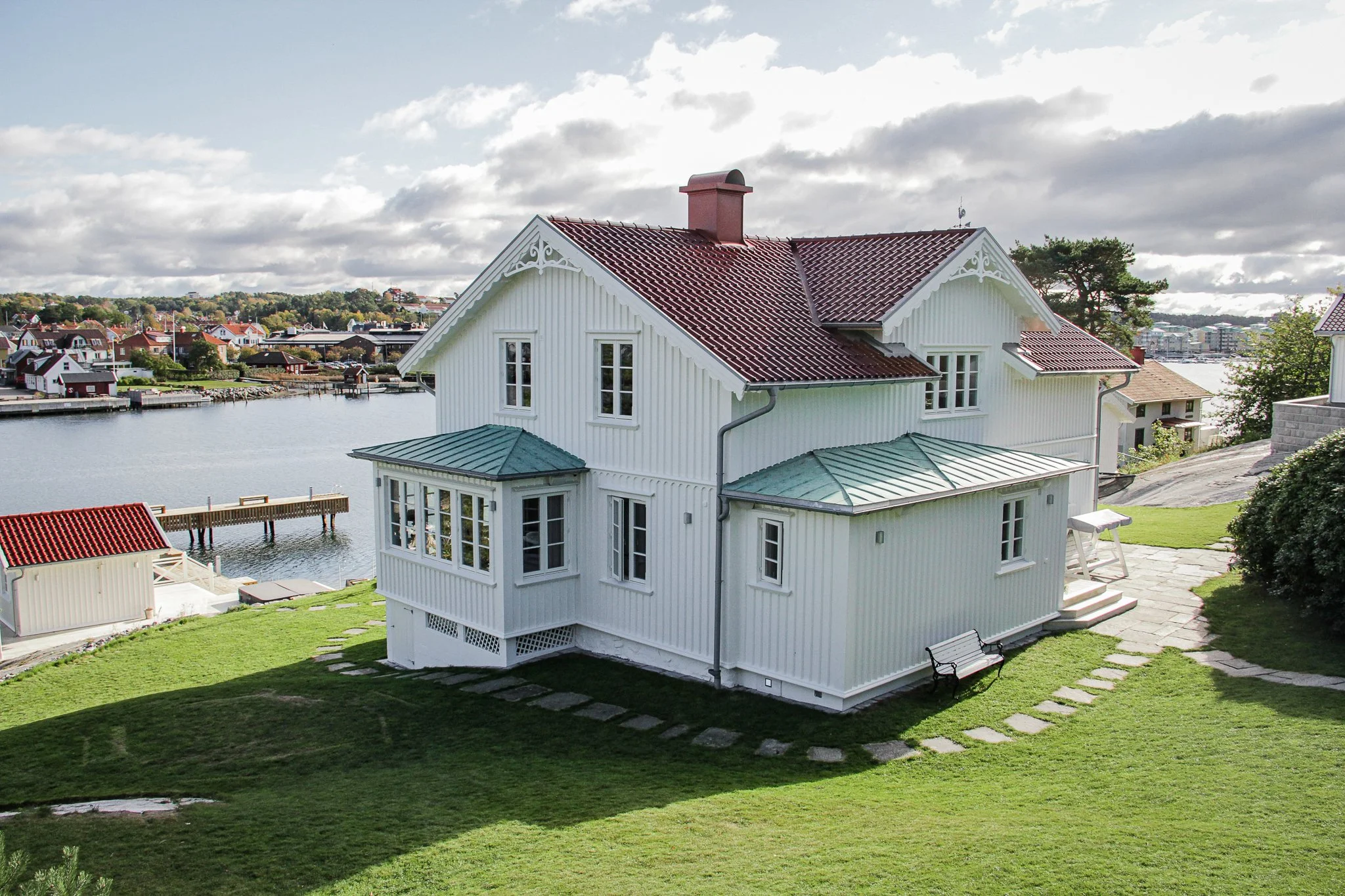 En vit villa vid vattnet med rött tak, gröna gräsmattor, och brygga. Flera hus i bakgrunden.