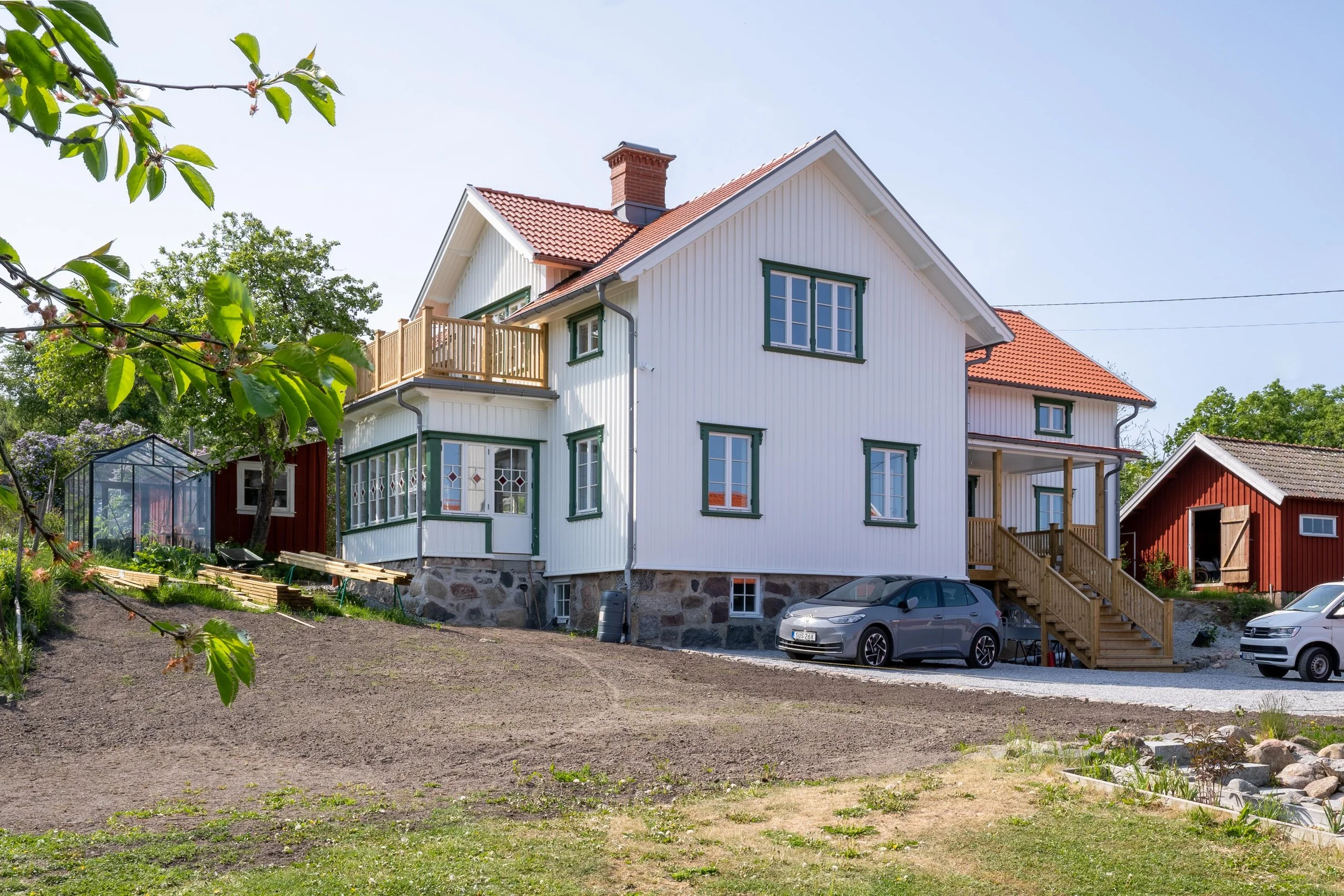 En vit trävilla med rött tegeltak och grönmålade fönsterkarmar, omgiven av träd och gräsmatta. En trappa leder upp till entrén, och en bil står parkerad framför huset. I bakgrunden finns en röd byggnad och ett växthus.