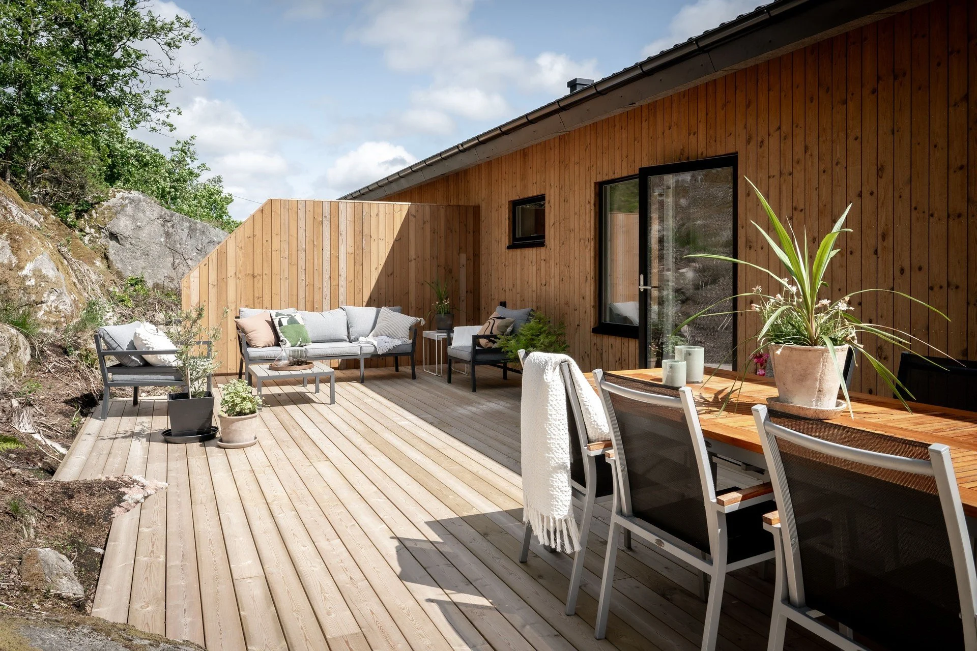 En uteplats med trädäck, soffgrupp och matbord, omgiven av träväggar och stenig terräng. Solig dag med gröna växter på bordet.