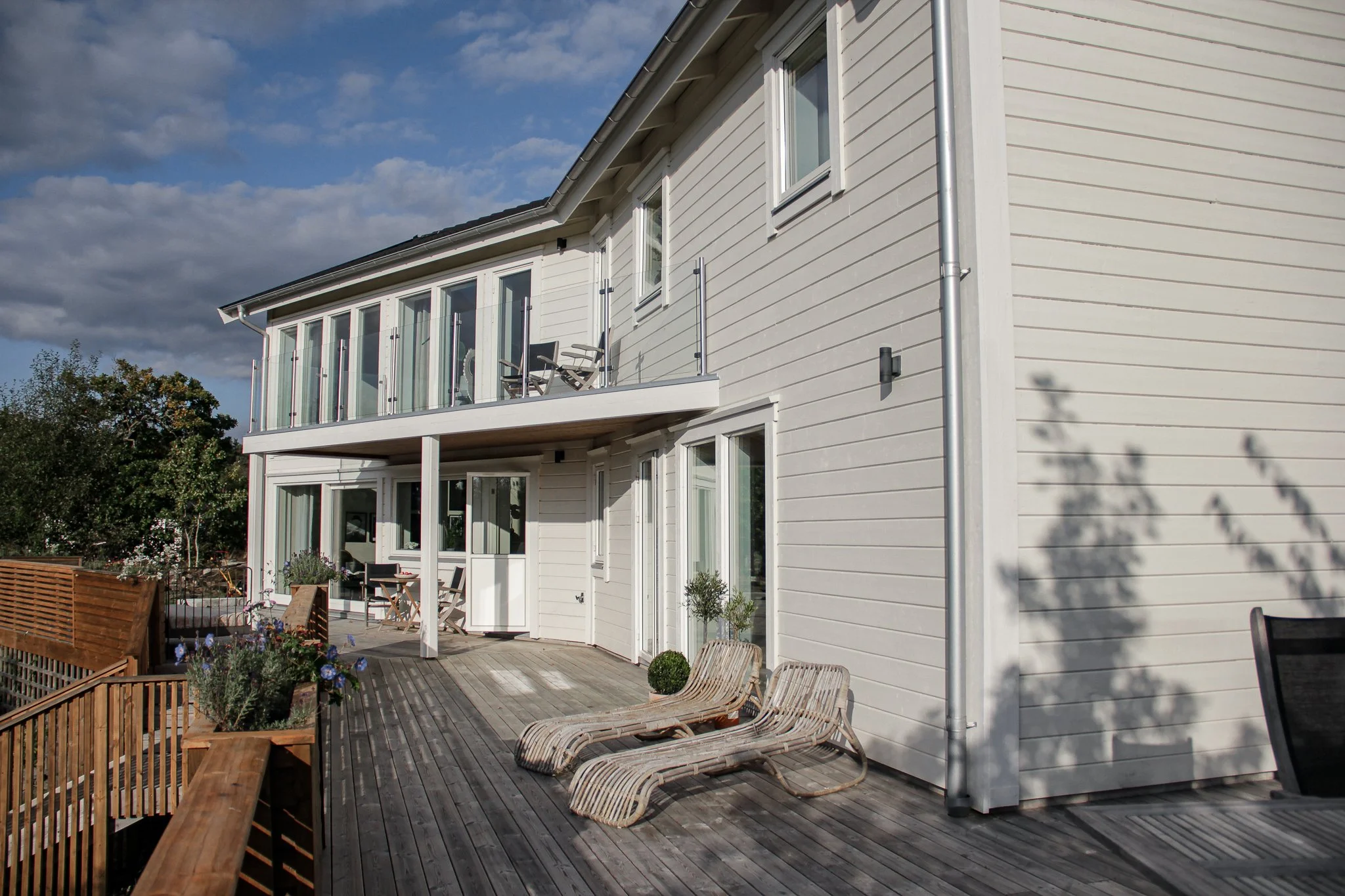 Modernt tvåvåningshus med träfasad och solig altan med solstolar.