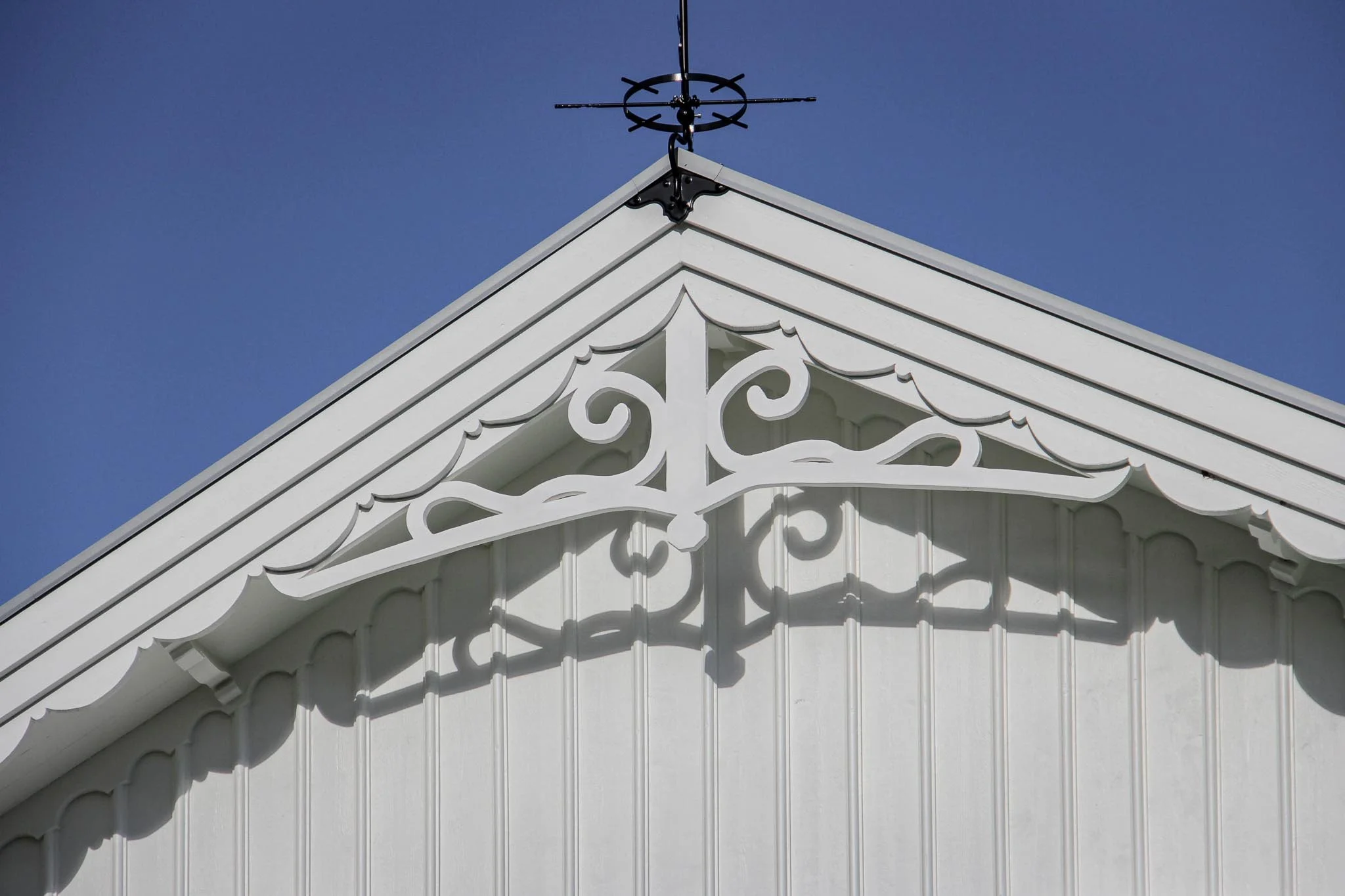 Vit träfasad med dekorativt snickeri och en väderflöjel mot en blå himmel.