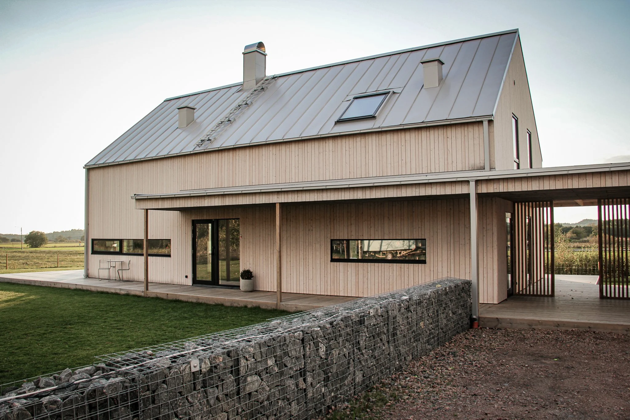 Modern träbyggnad med metalltak och stenmur framför, omgiven av grönska.
