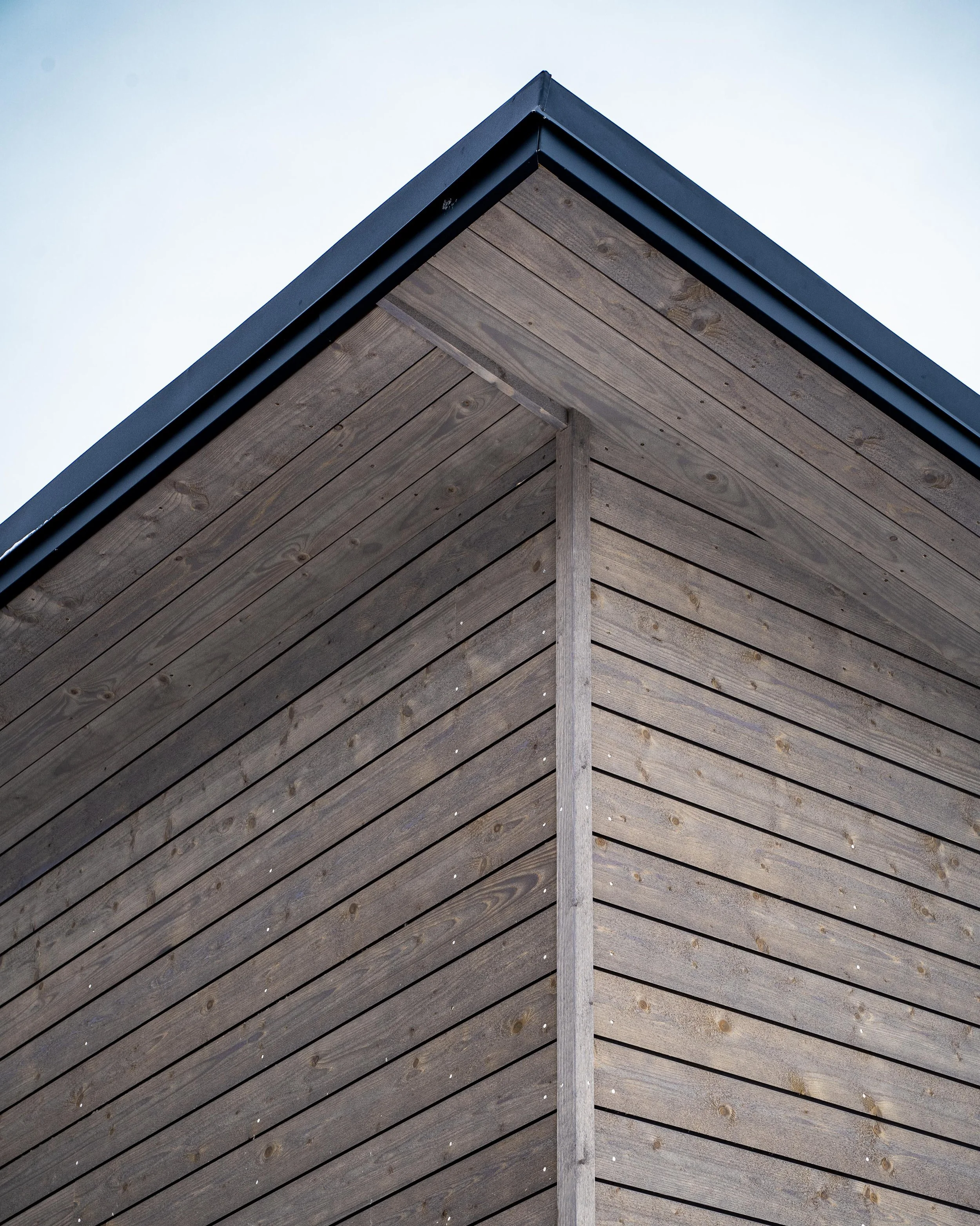Hörnet av ett modernt hus med träpaneler och ett platt tak. Ljusa träplankor ger ett stilrent utseende. Taken är svart och korsar de vertikala och horisontella linjerna av väggpanelen. Grå himmel i bakgrunden.