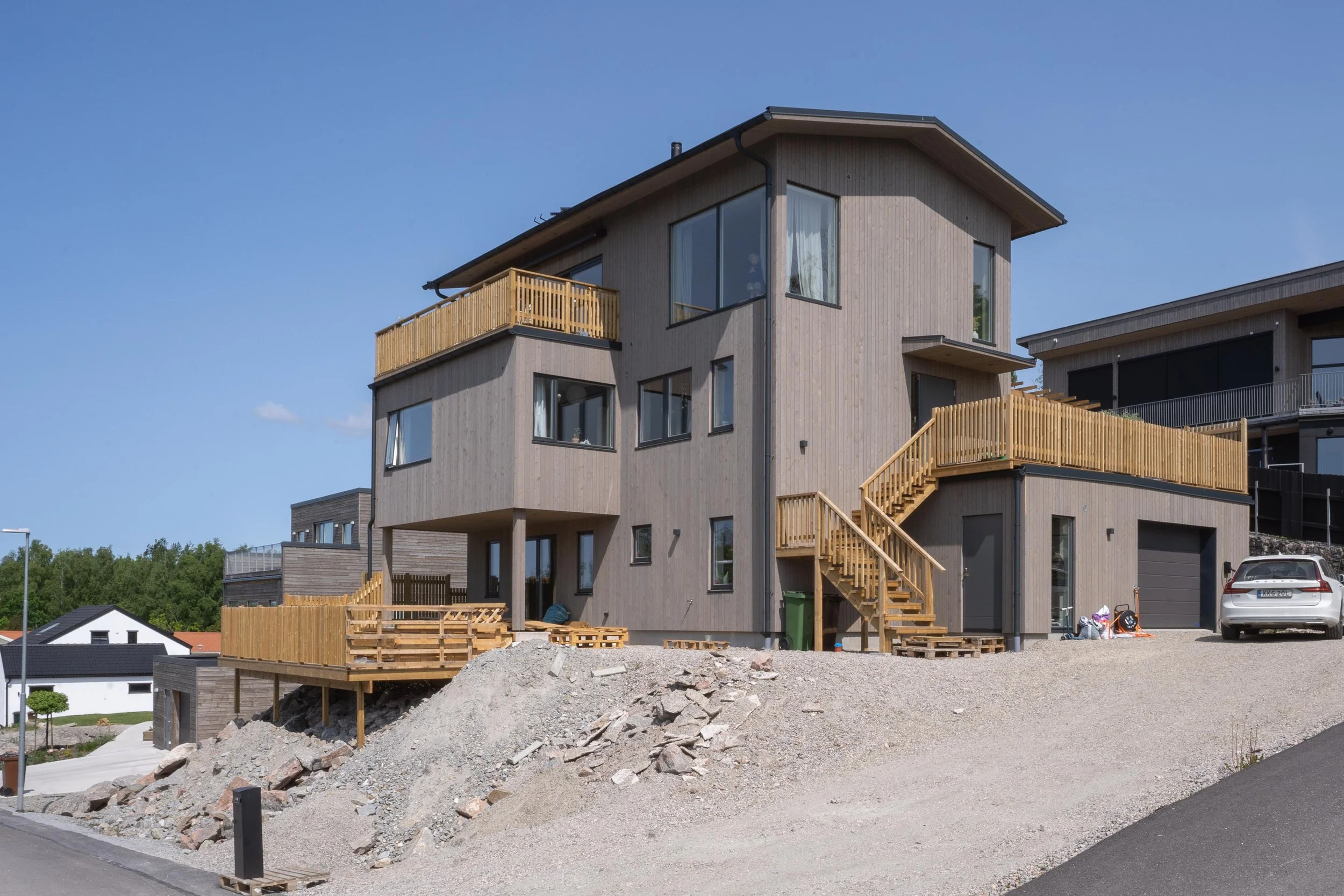 Modern träklädd villa med flera våningar och terrasser, vit bil parkerad framför, klart väder.