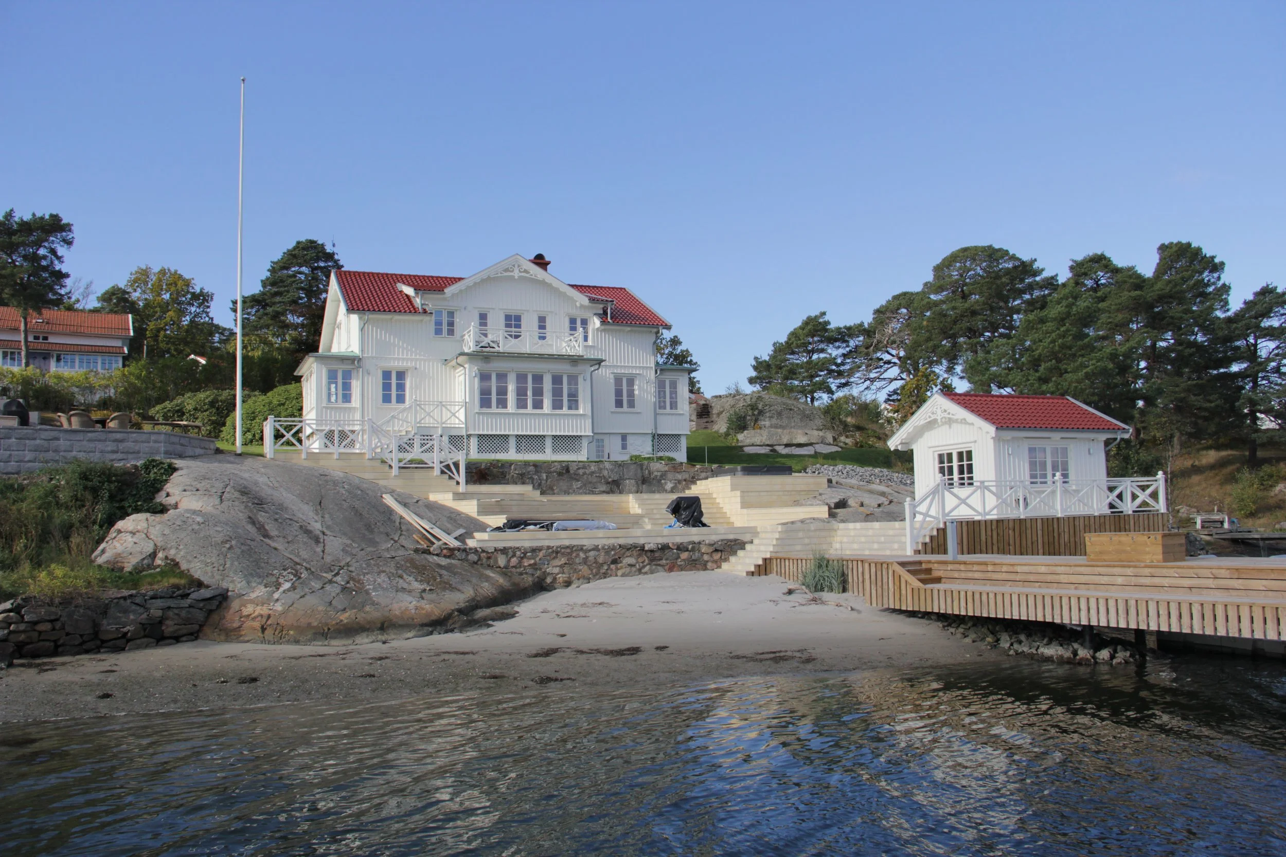 Vit villa vid vattnet med rött tak och brygga, omgiven av träd.