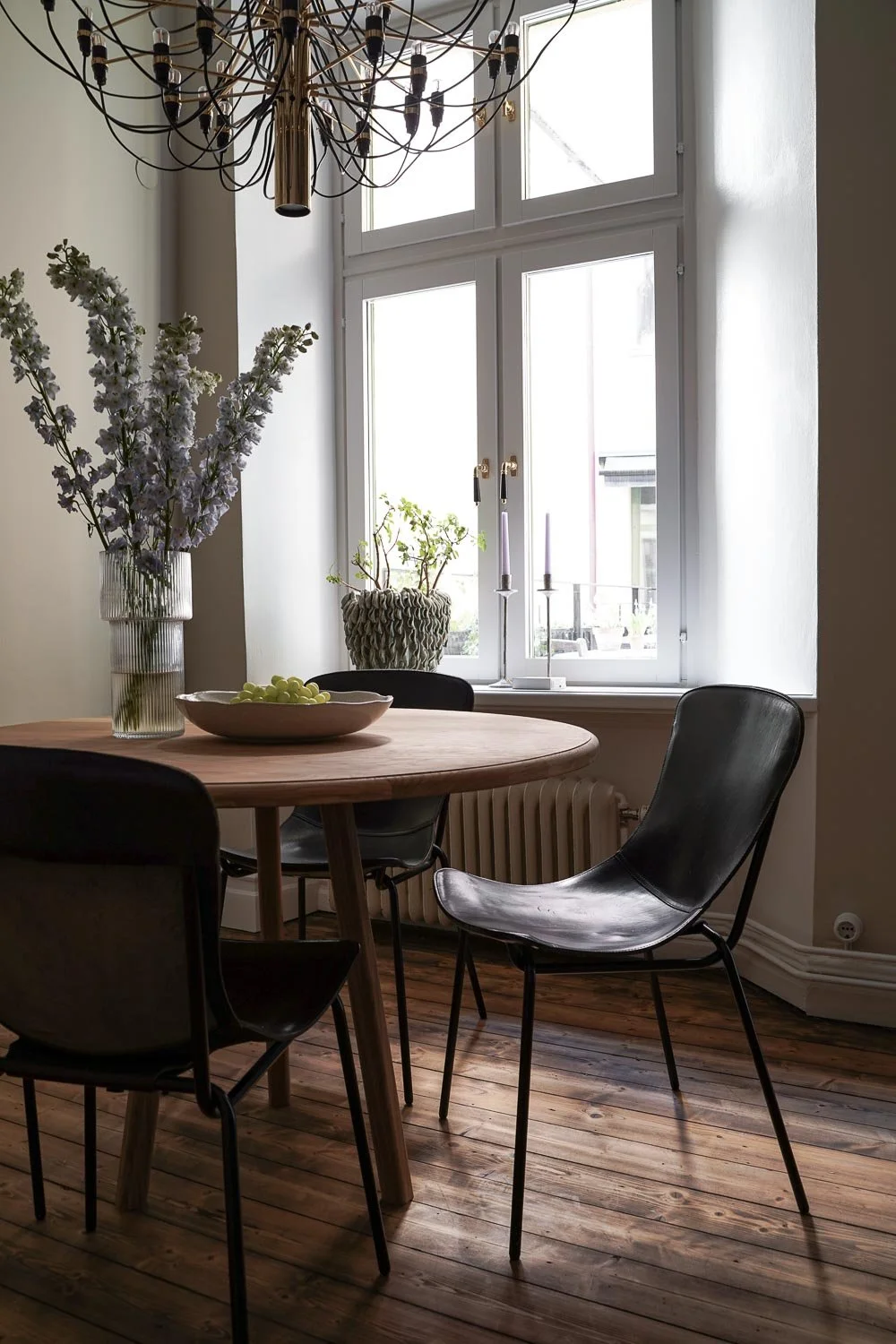 Matbord av trä med svarta stolar, vas med blommor och fruktskål framför ett fönster med stearinljus, ljuskrona i taket, trägolv. O Lind AB.
