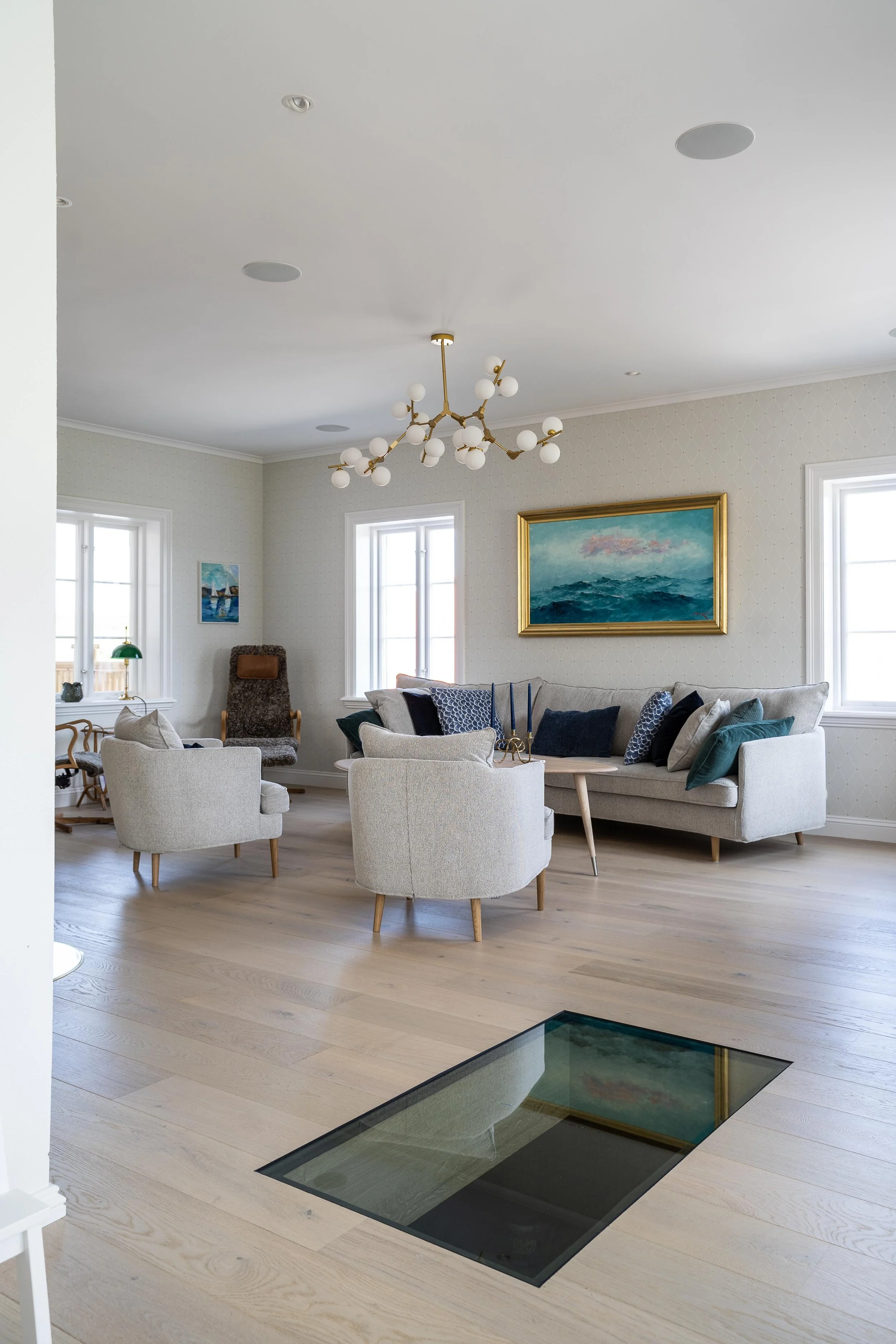 Modernt vardagsrum med en ljuskrona i taket, en soffa, fåtöljer och kuddar, samt ett glasgolv. Tavlor hänger på väggarna. O Lind AB.