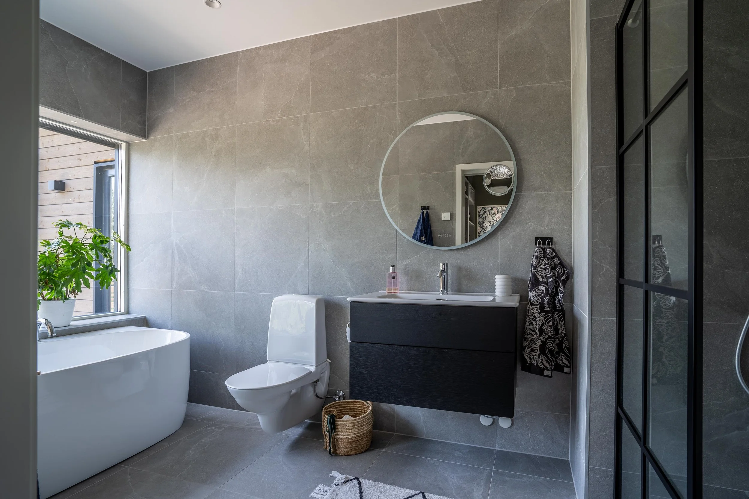 Modernt badrum med grå väggar, fristående badkar, toalett, spegel. En växt i kruka står vid fönstret. O Lind AB.