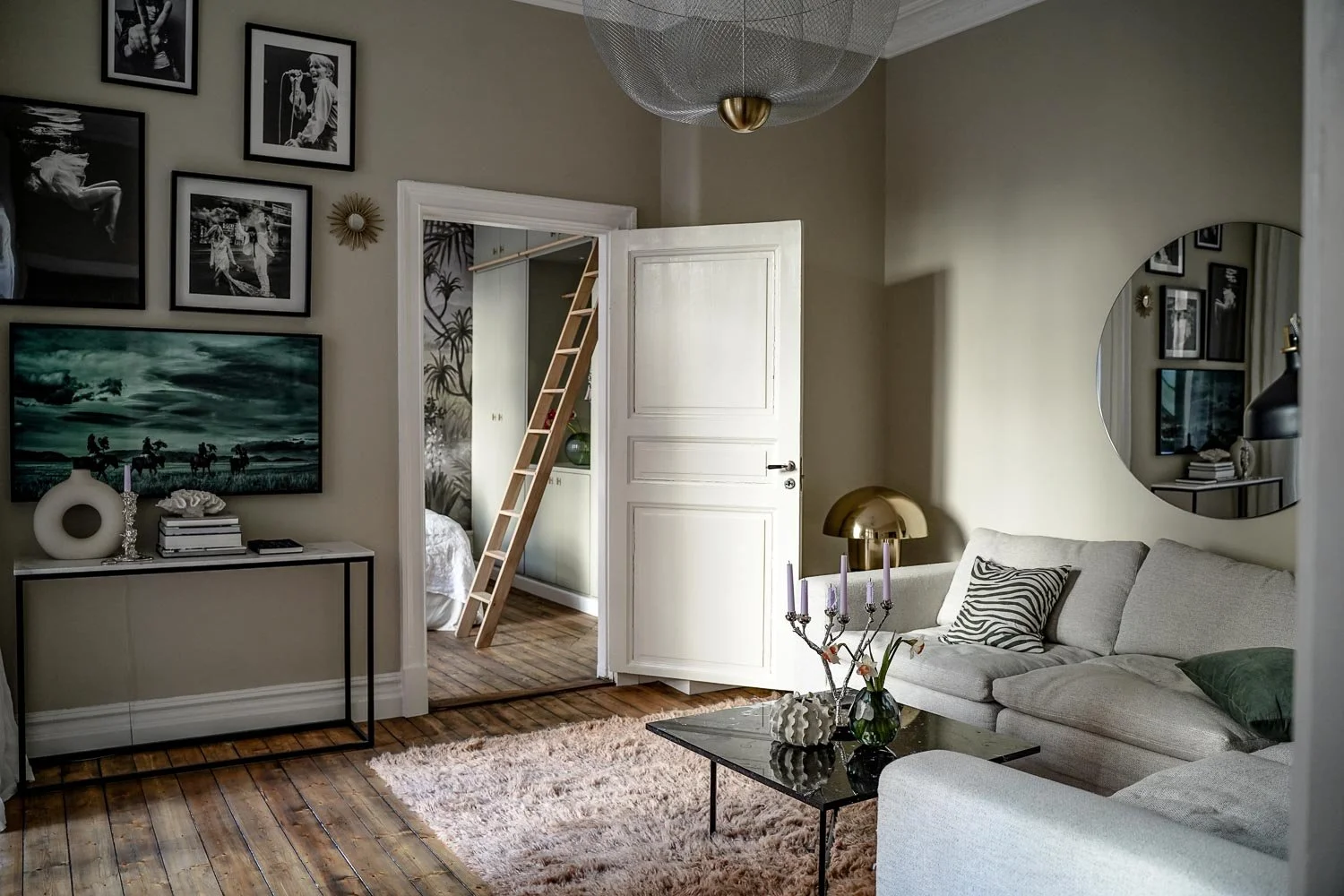 Elegant vardagsrum med ljusgrå soffa, runda spegeln på väggen, svart marmorbord, dekorativa kuddar, och väggkonst. Öppen dörr leder till sovrum, trästege synlig. Mjuk matta på trägolv, stilfull belysning och ljusstakar. O Lind AB.