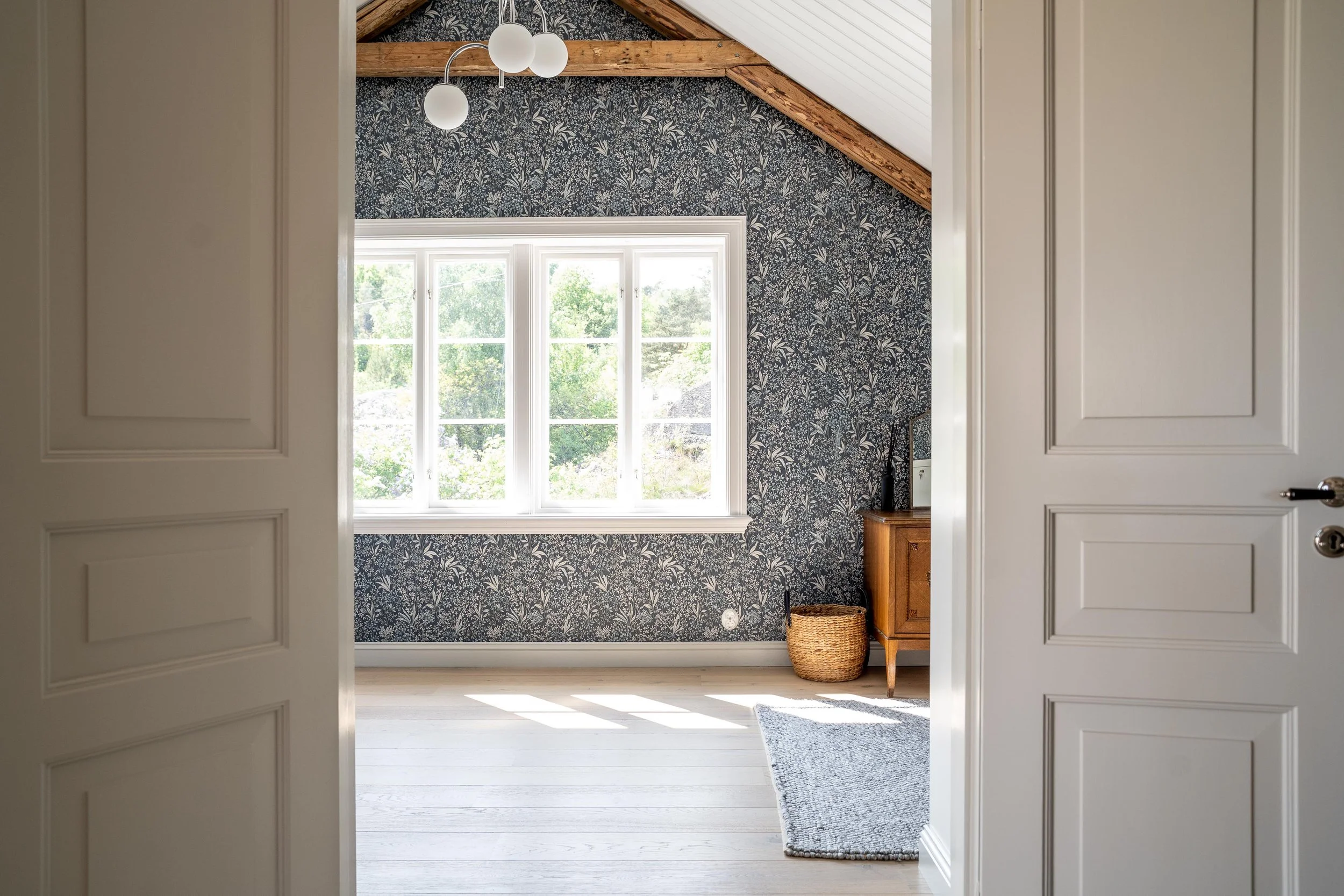 Ett rum med en tapetserad vägg i blommigt mönster, stora fönster med utsikt över grönska, en hängande taklampa, en flätad korg och en träbyrå. Golvet är ljust trä och det finns en grå matta. Taken är vita med synliga träbalkar. O Lind AB.